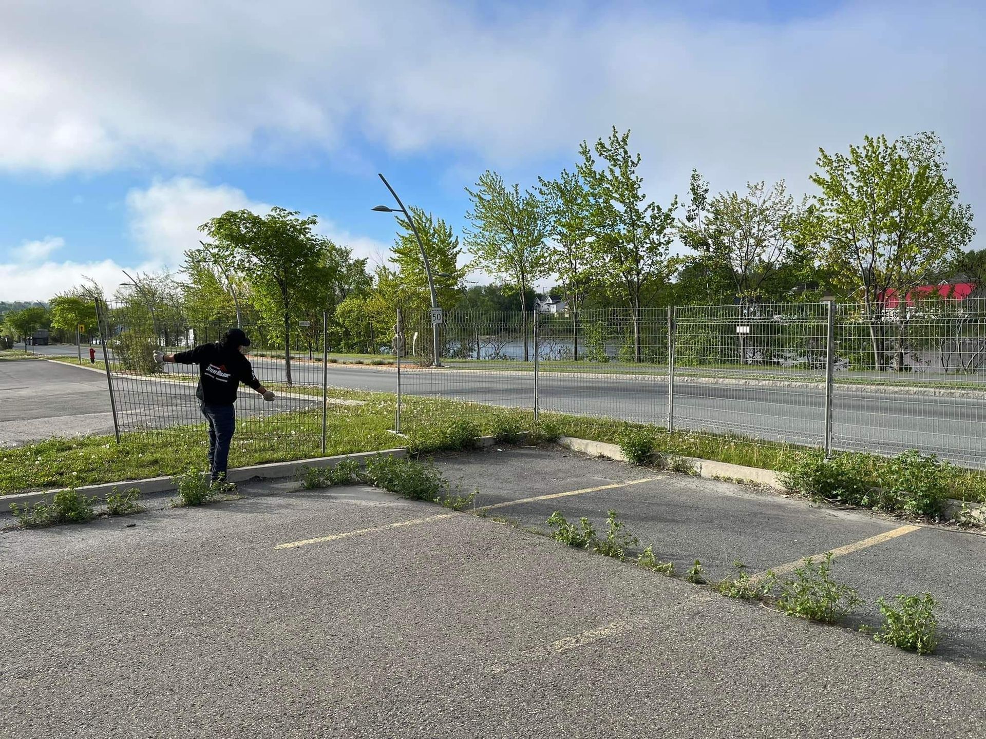 Une personne marche sur un parking presque vide, avec de petits arbres et une clôture en arrière-plan, par temps nuageux.