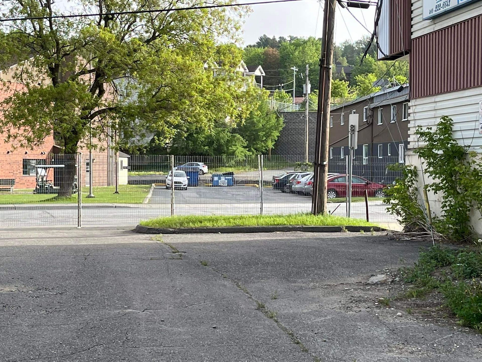 Une ruelle avec des voitures garées, une clôture, un bâtiment et un arbre par une journée ensoleillée.