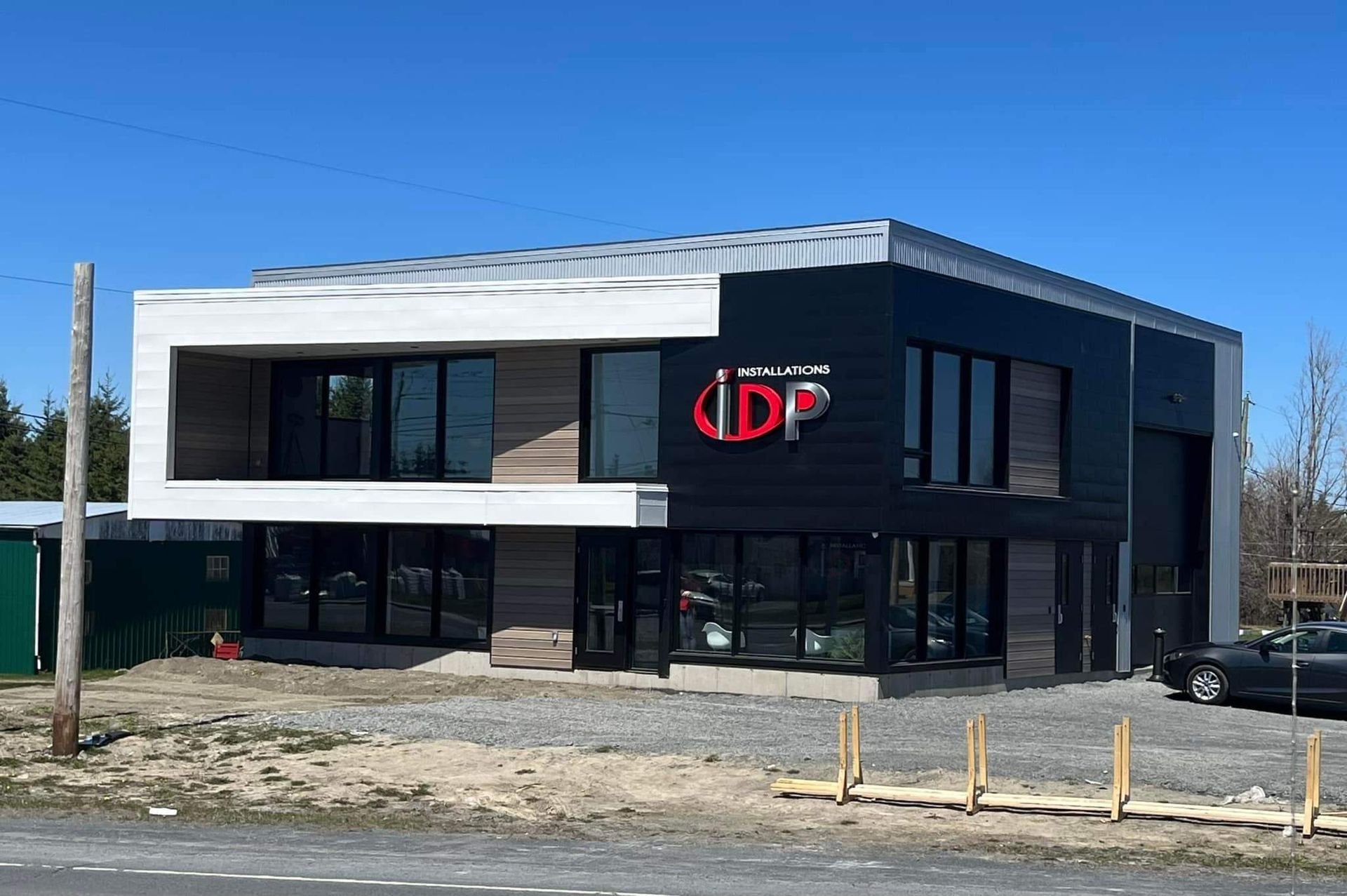 Bâtiment moderne de deux étages avec logo « IDP » rouge, façade noire et blanche et grandes fenêtres. Gravier et voiture visibles.