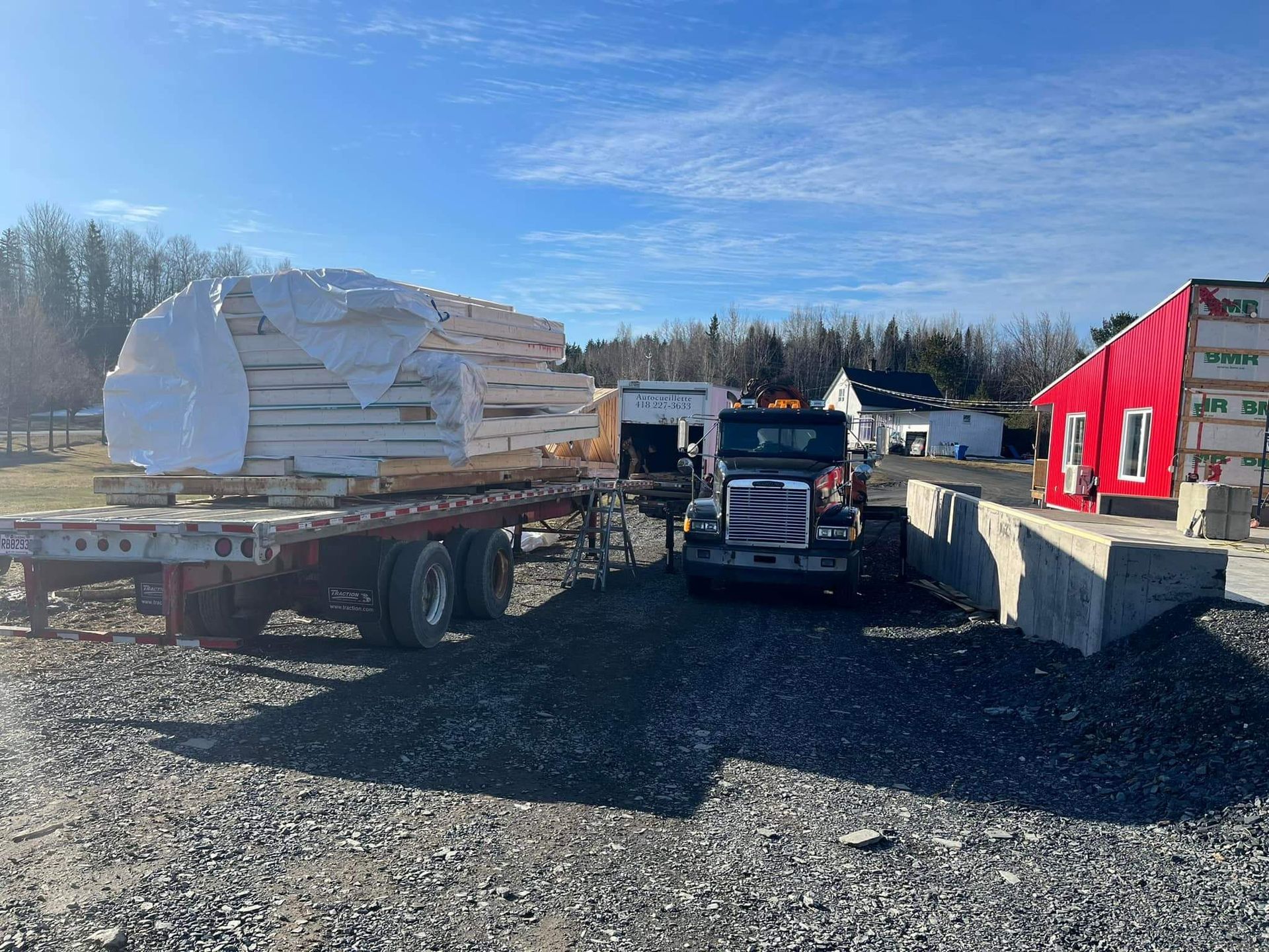 Un camion avec une remorque à plateau transporte une importante cargaison de matériaux emballés en blanc sur un terrain gravillonné à côté d'un bâtiment rouge.