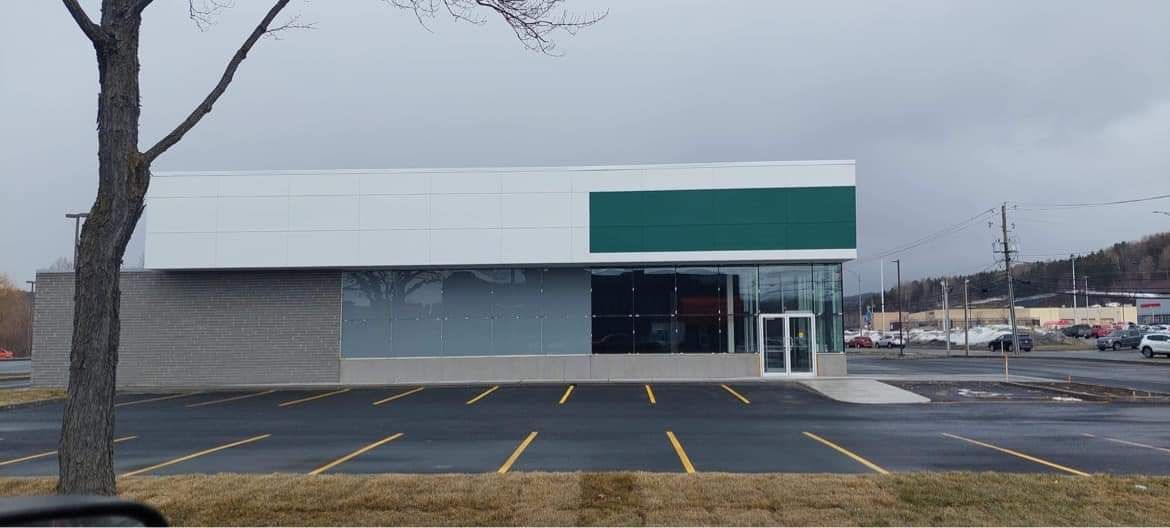 Extérieur d'un bâtiment commercial avec une façade blanche et verte, des fenêtres en verre et un parking pavé.
