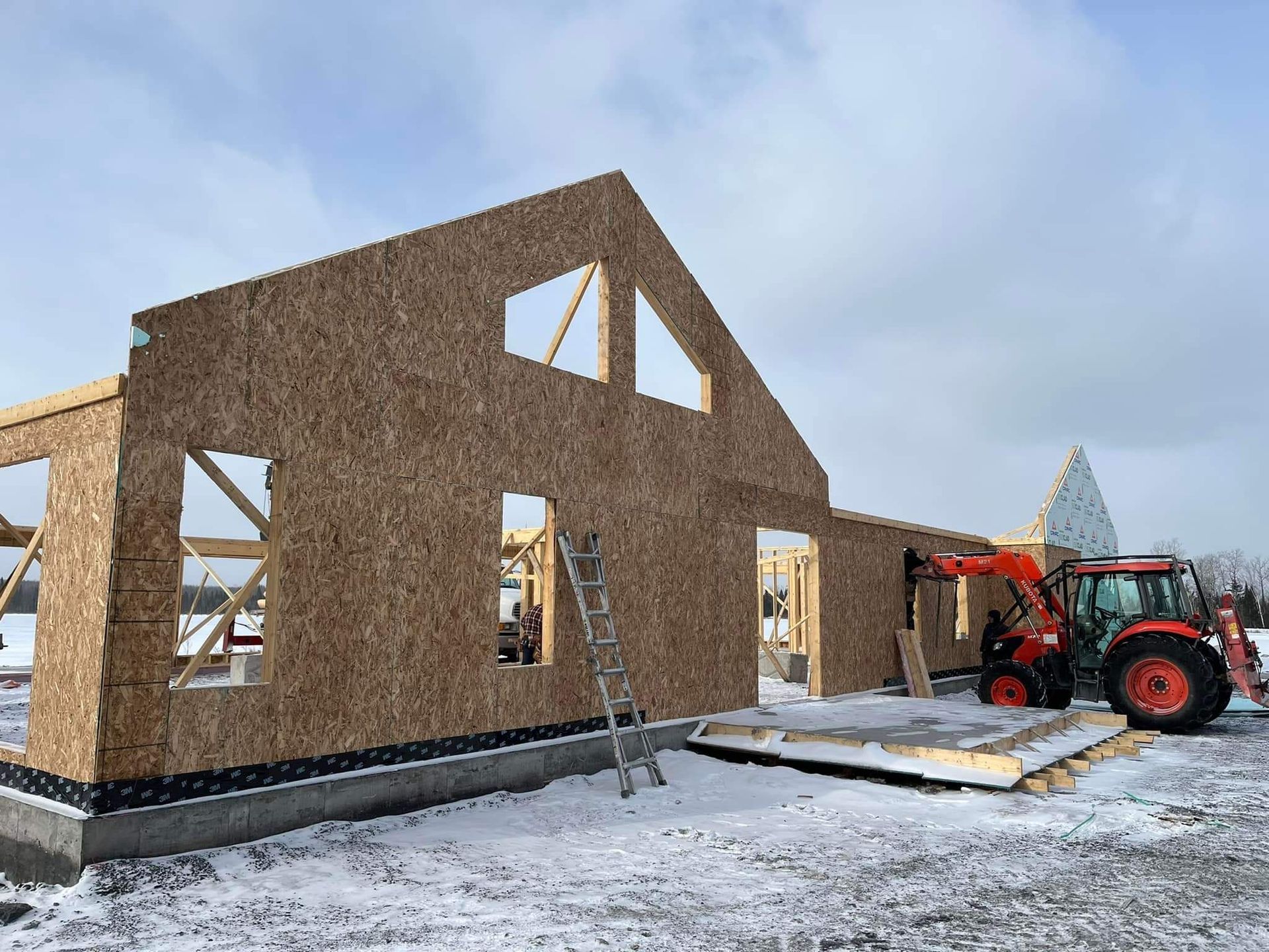 Chantier de construction avec une maison à ossature bois en construction ; tracteur orange garé à proximité, neige au sol.