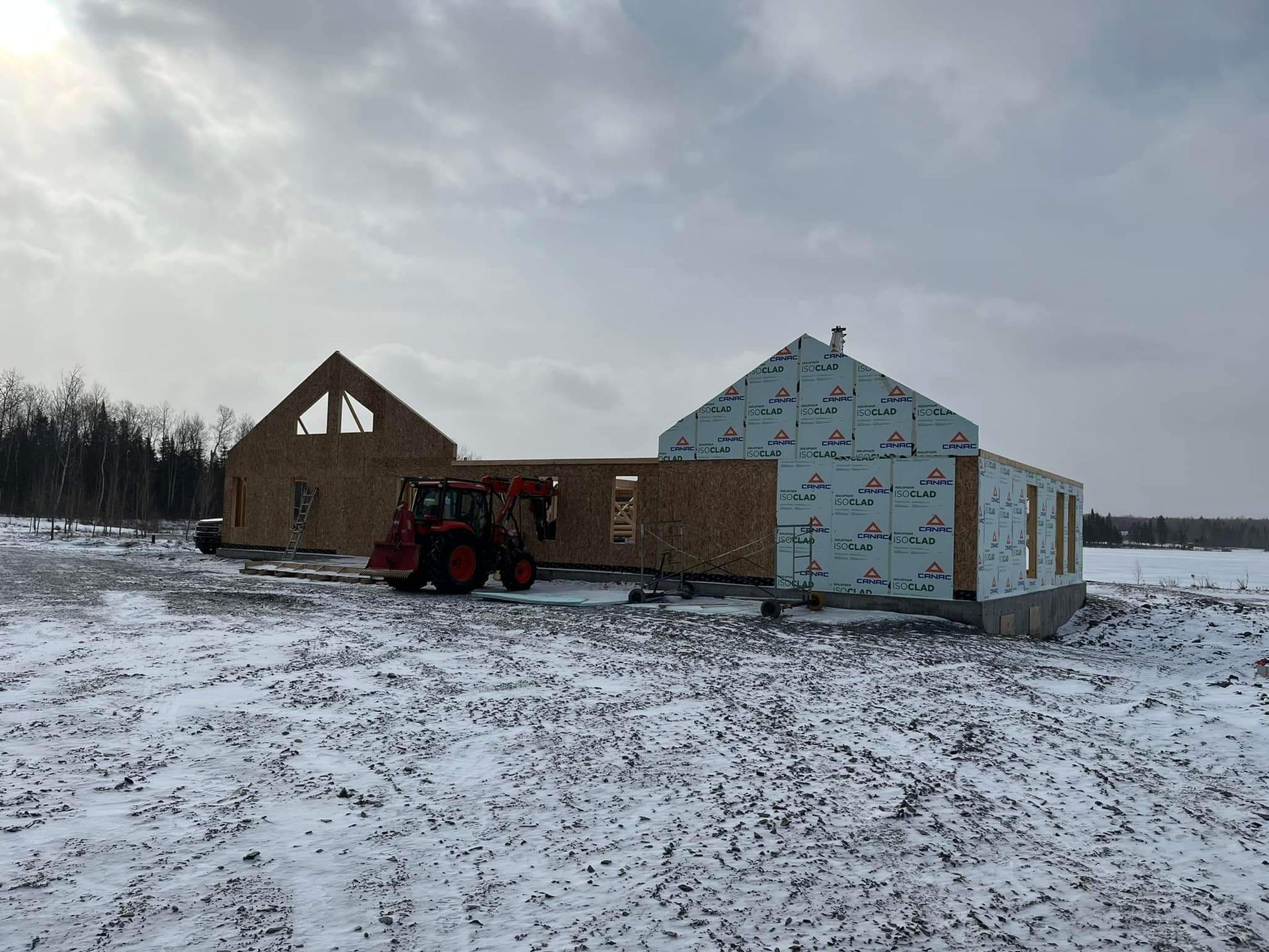 Chantier de construction d'une maison avec charpente et murs extérieurs dans un champ enneigé. Un tracteur est présent.