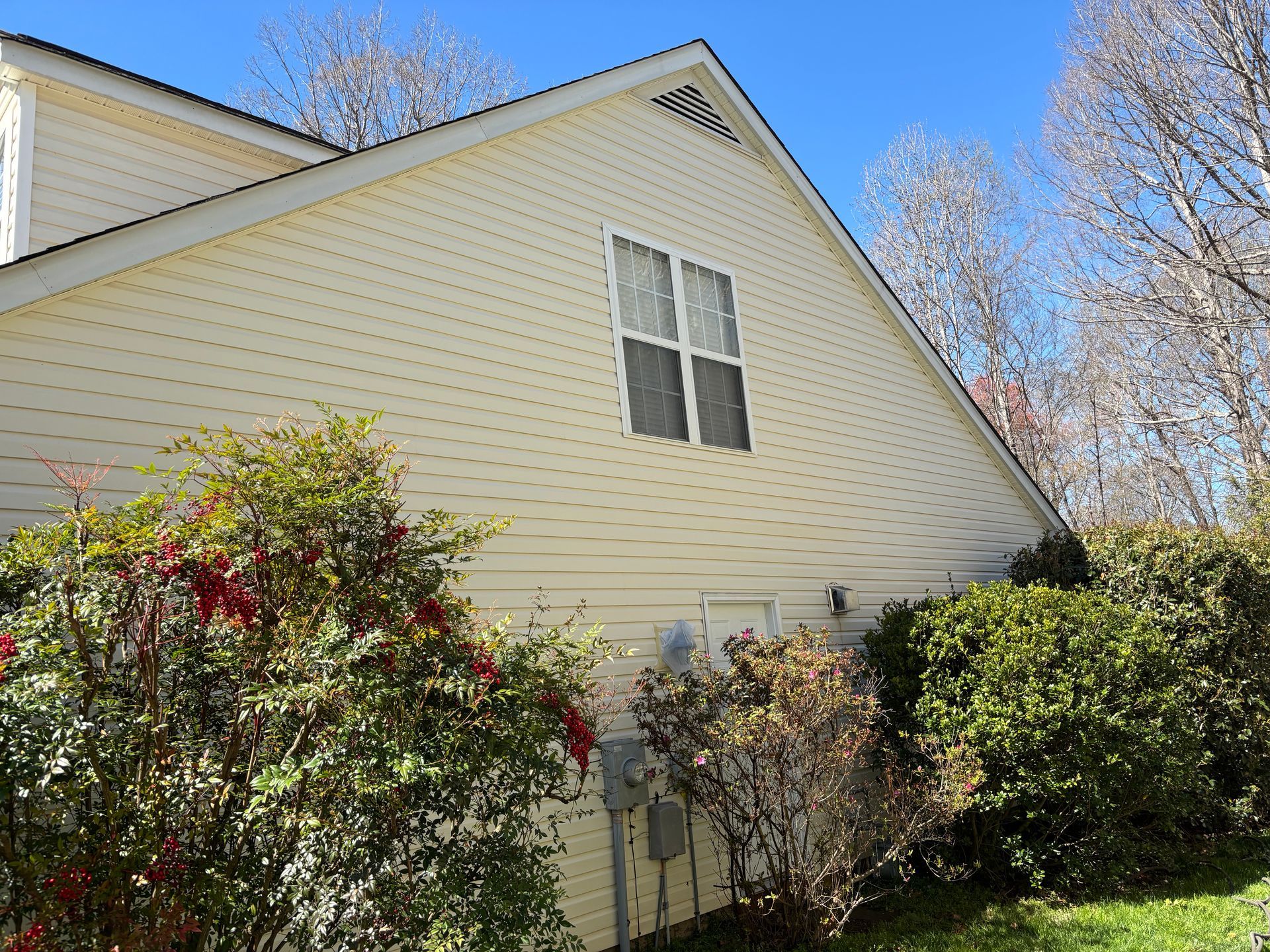 Windows Gutters LLC - Yellow-sided house with a small window in the gable and bushes in the foreground against a blue sky.