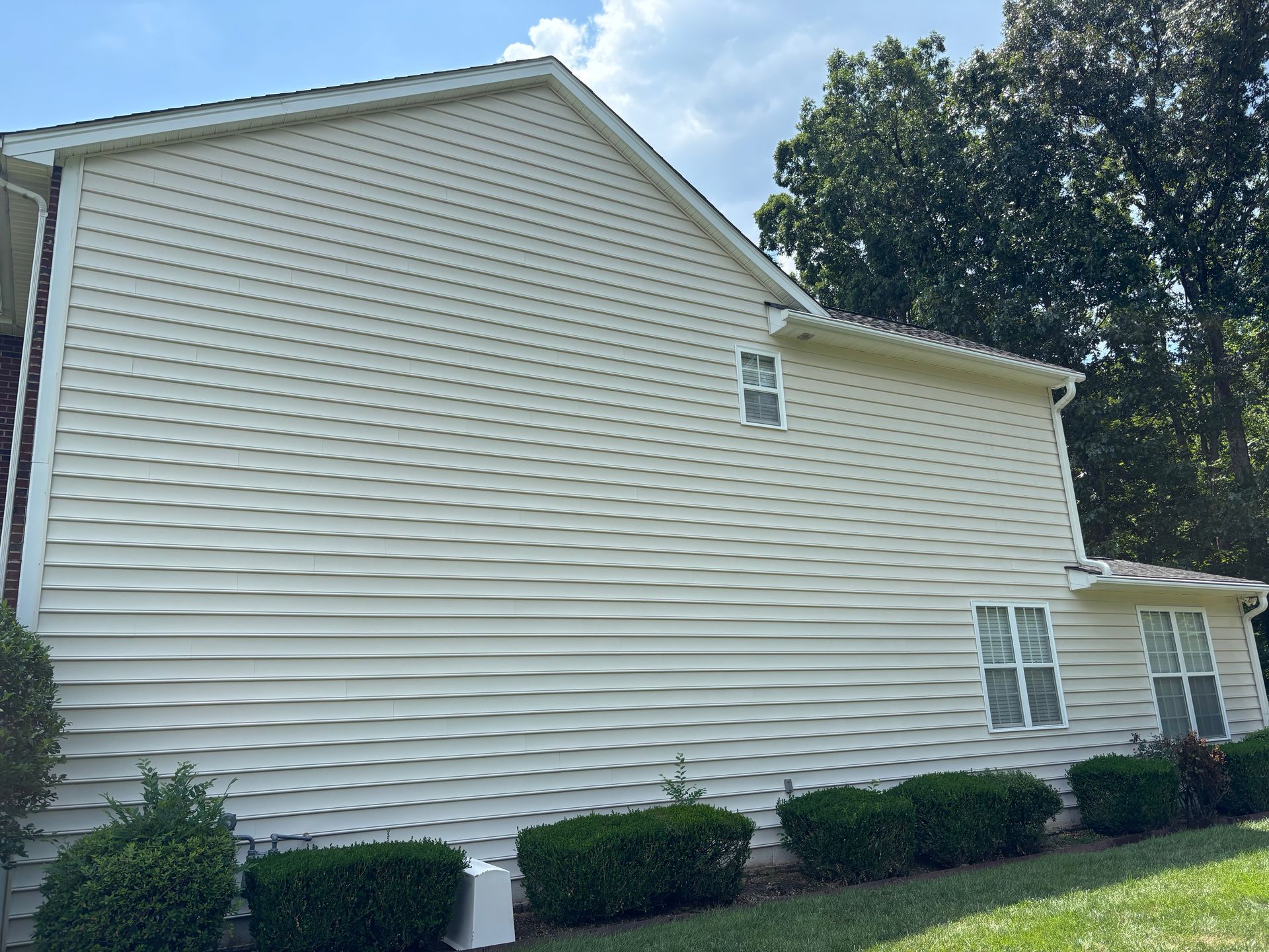 Windows Gutters LLC - Side of a beige house with white trim, windows, and green bushes in front. Blue sky.
