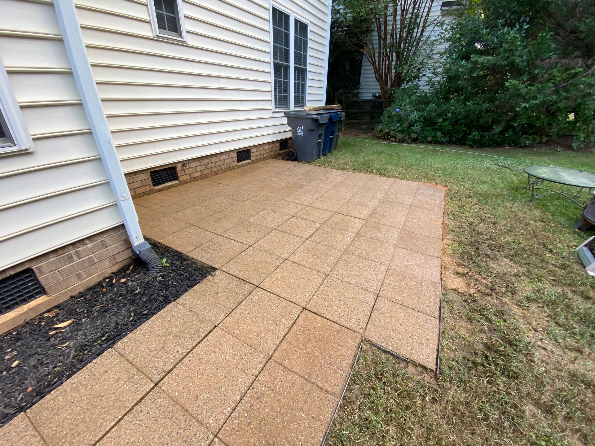 Windows Gutters LLC - A patio made of brown square tiles next to a house with cream siding and green grass.