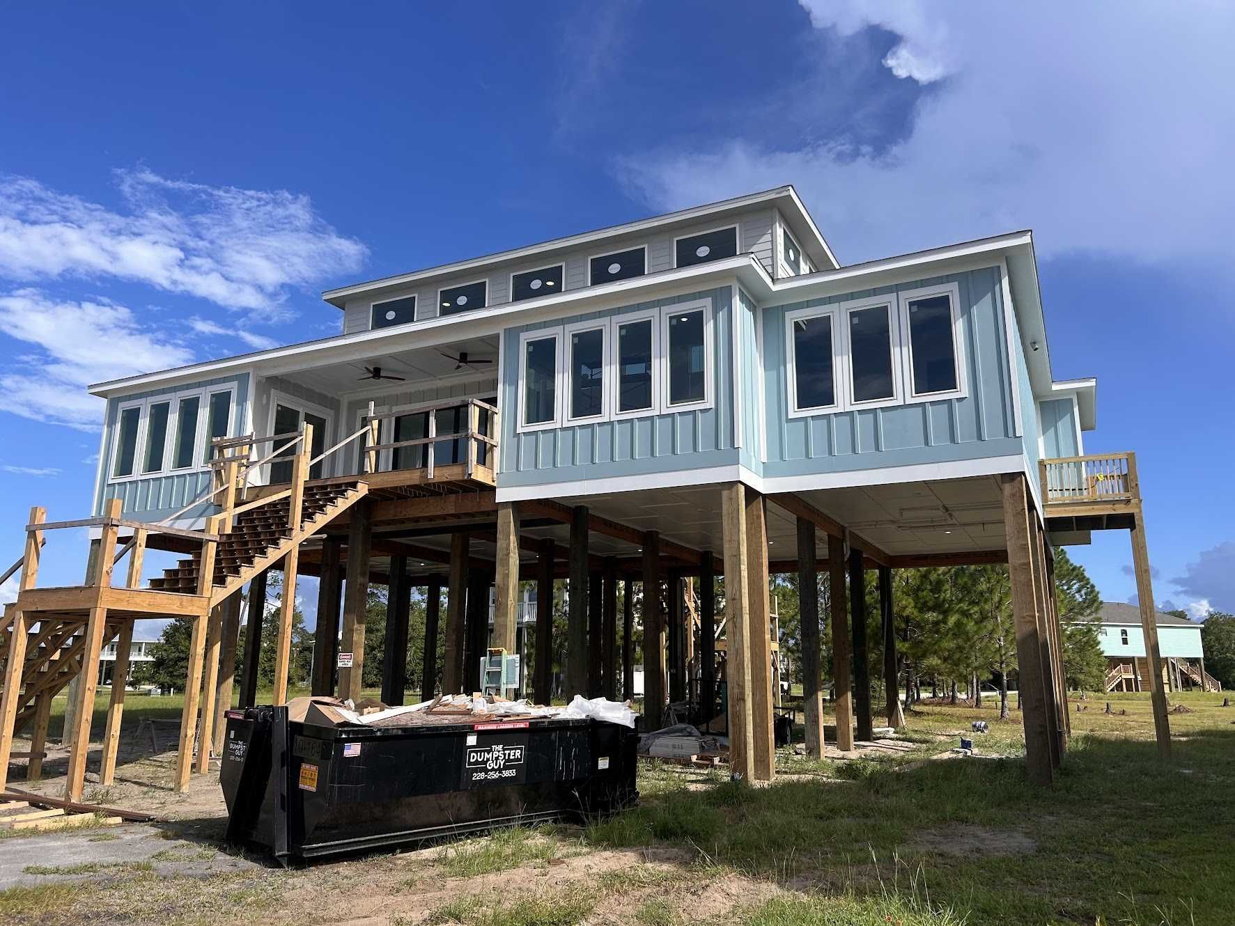 A light blue beach house under construction, elevated on wooden pilings, with a large dumpster in the foreground.