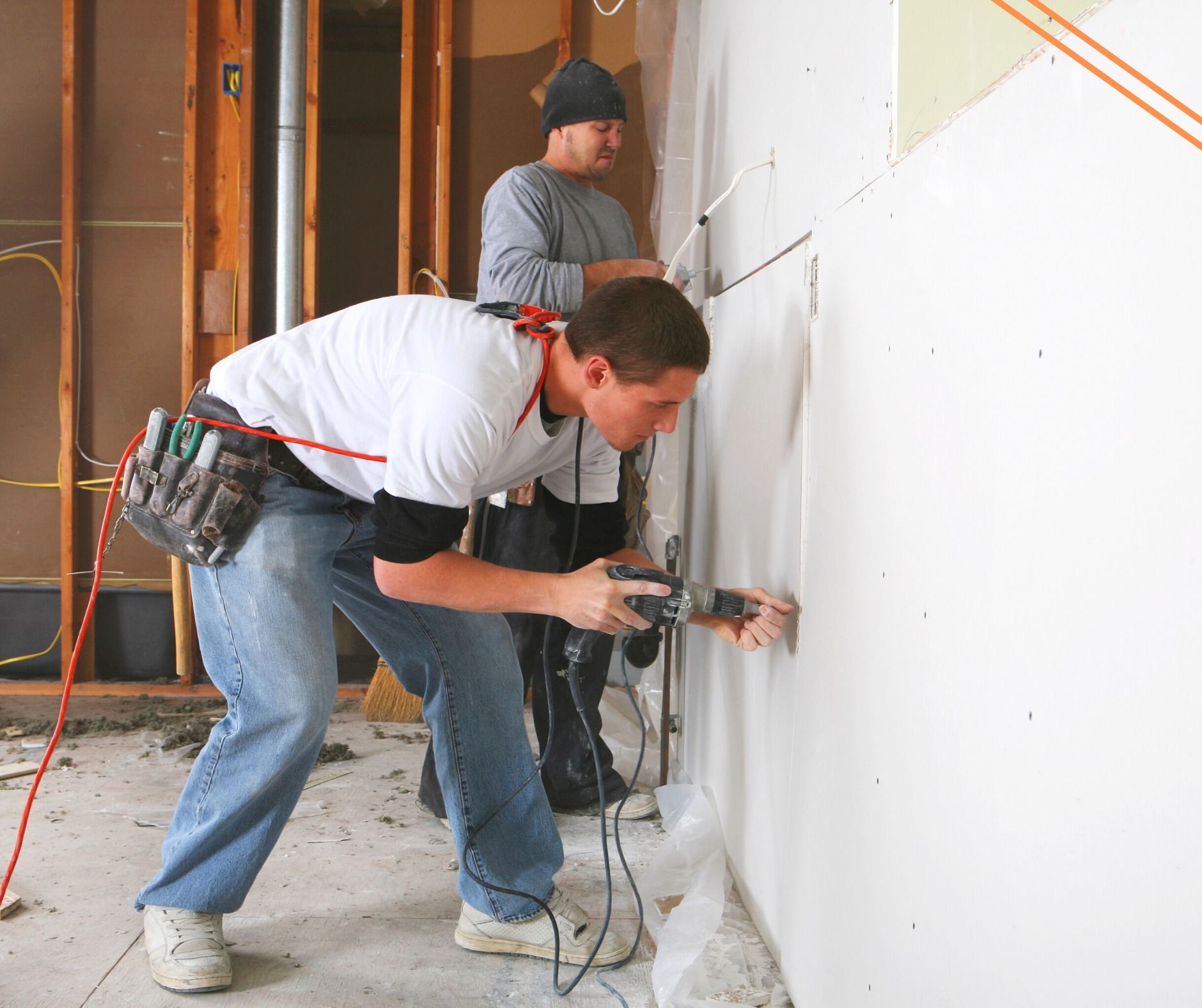 Two construction workers installing drywall on a wall. One uses a power tool while the other watches in a room with exposed studs.
