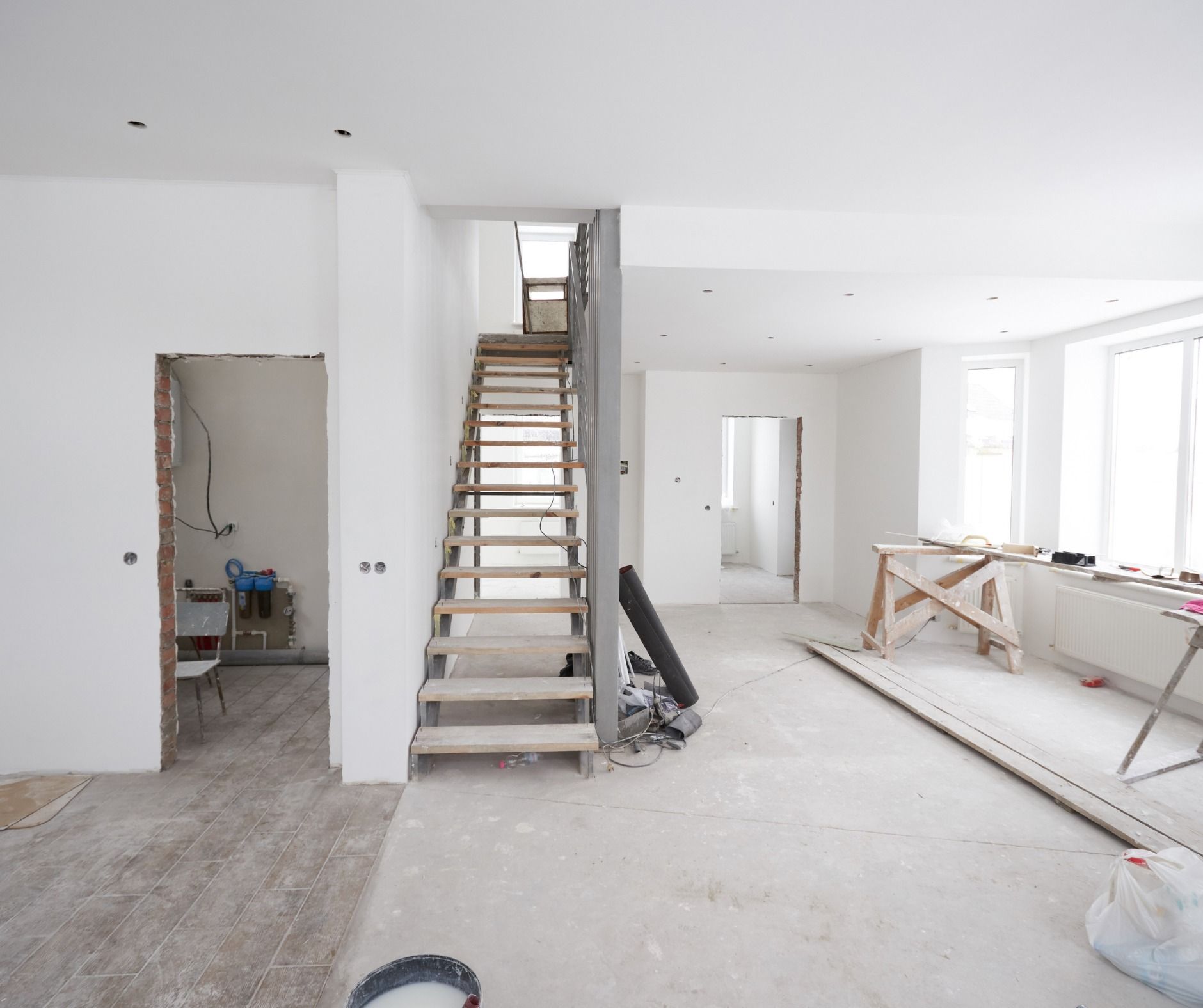 Interior view of a home under construction. Staircase, open doorways, and construction materials are visible on the unfinished floors.