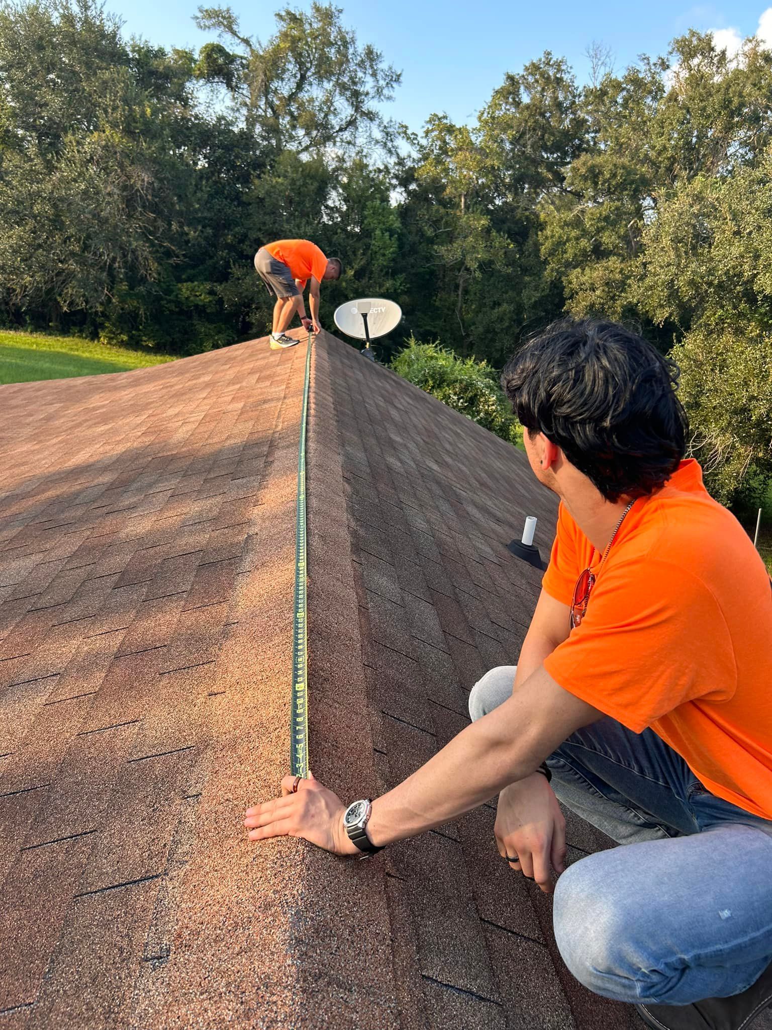 Two roofers in orange shirts measure a roof ridge, one standing, one kneeling. A satellite dish is visible.