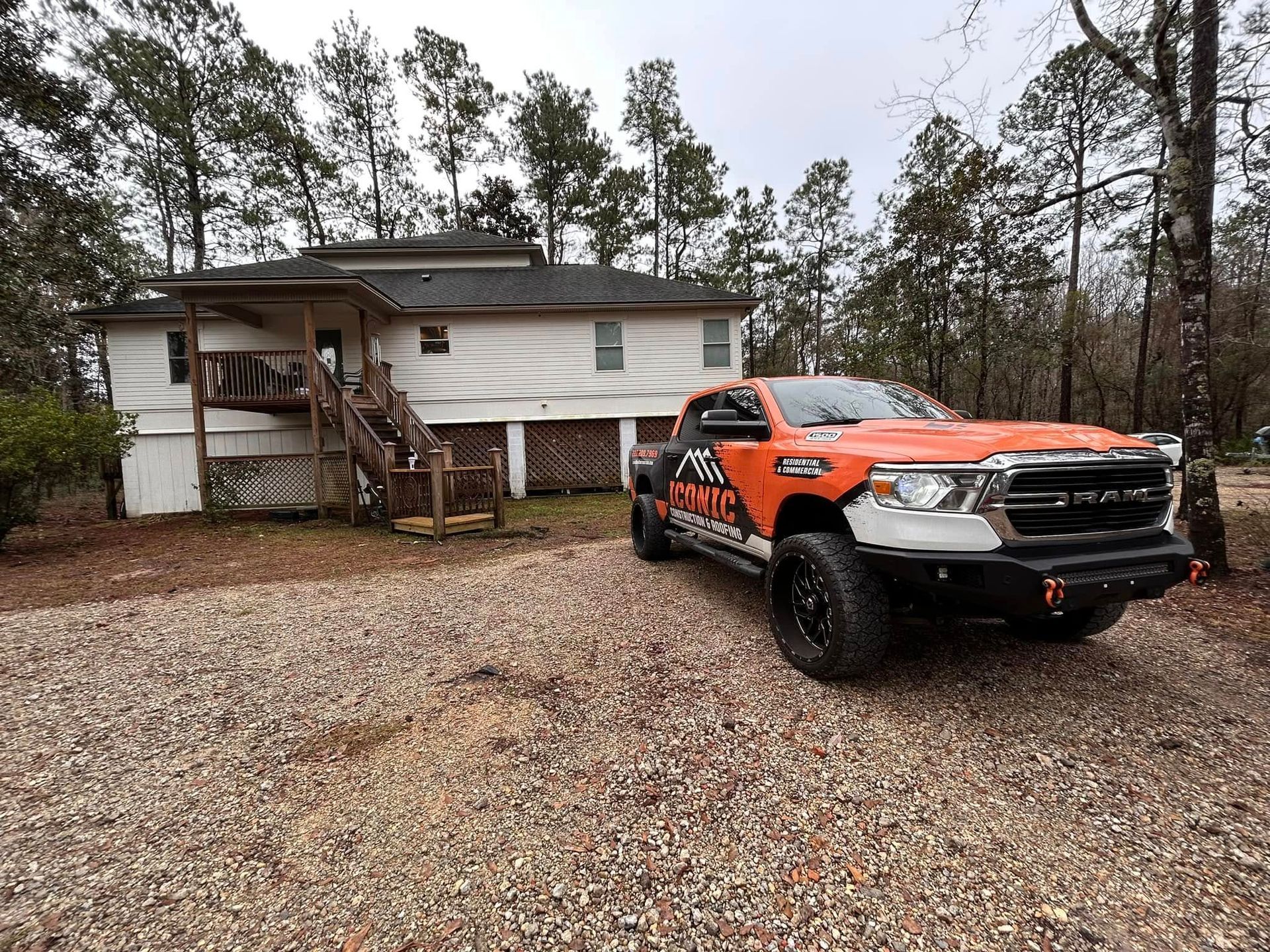 Orange and black truck parked in front of a two-story, white house with brown steps and a gravel driveway, surrounded by trees.