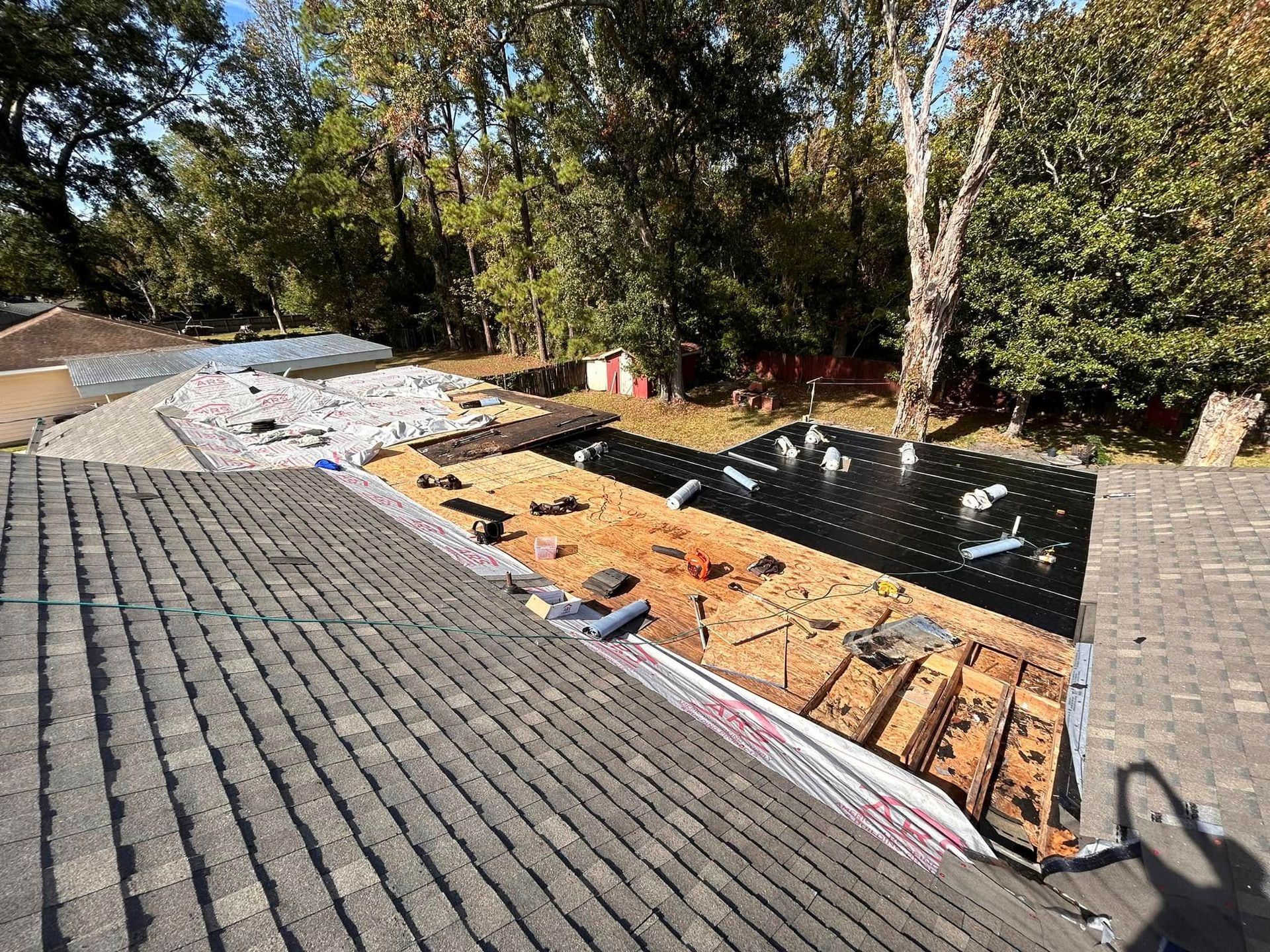 Rooftop under construction; old shingles and exposed underlayment with tarp and lumber visible; green trees in the background.