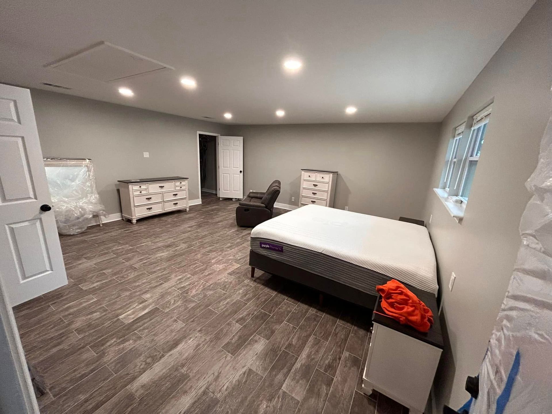 Spacious bedroom with grey walls, wood-look flooring, a bed, dressers, and a recliner. There's a door and a window.