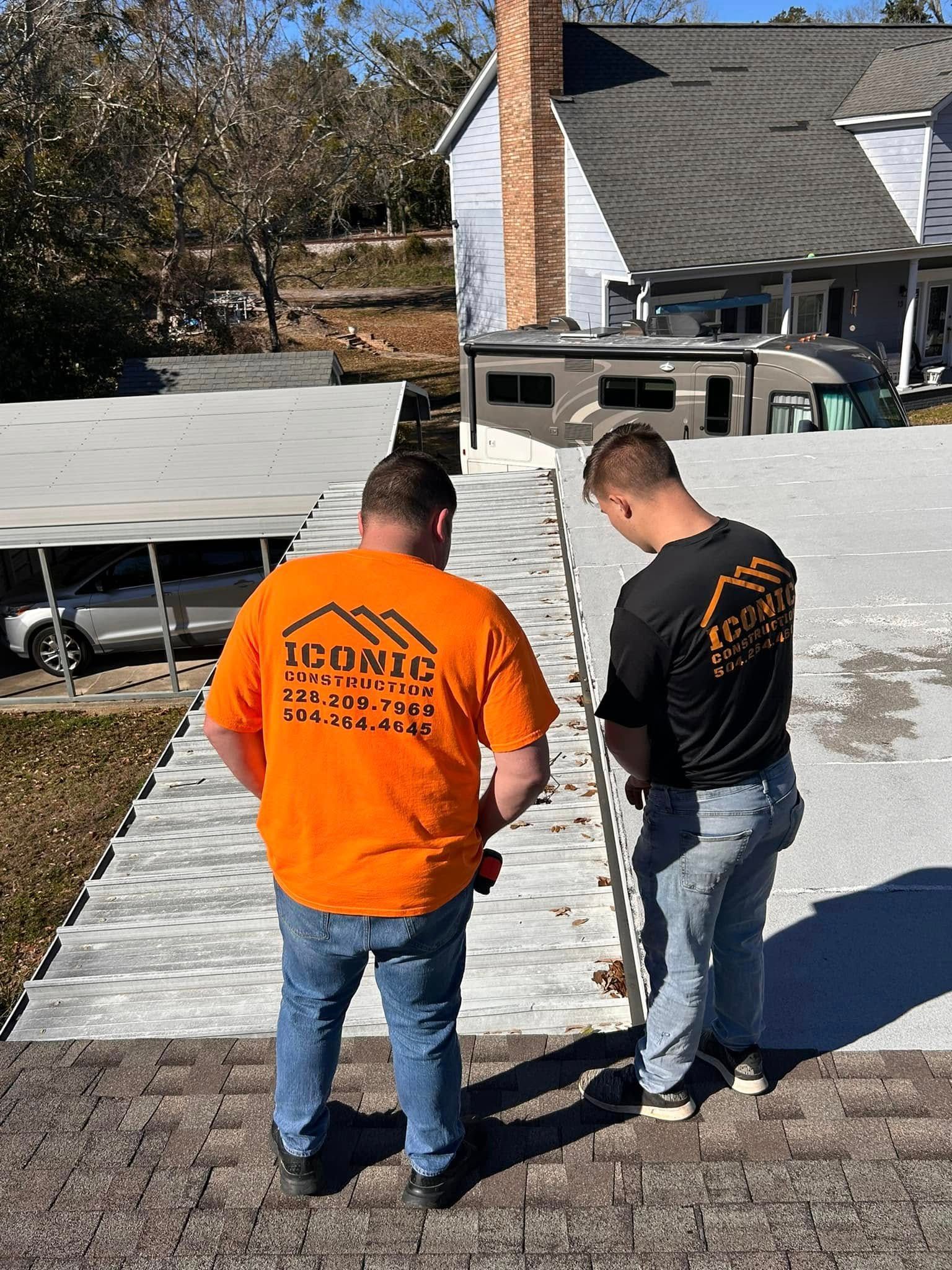 Two roofers wearing orange and black shirts are working on a roof. The roof is light gray, and a house is in the background.