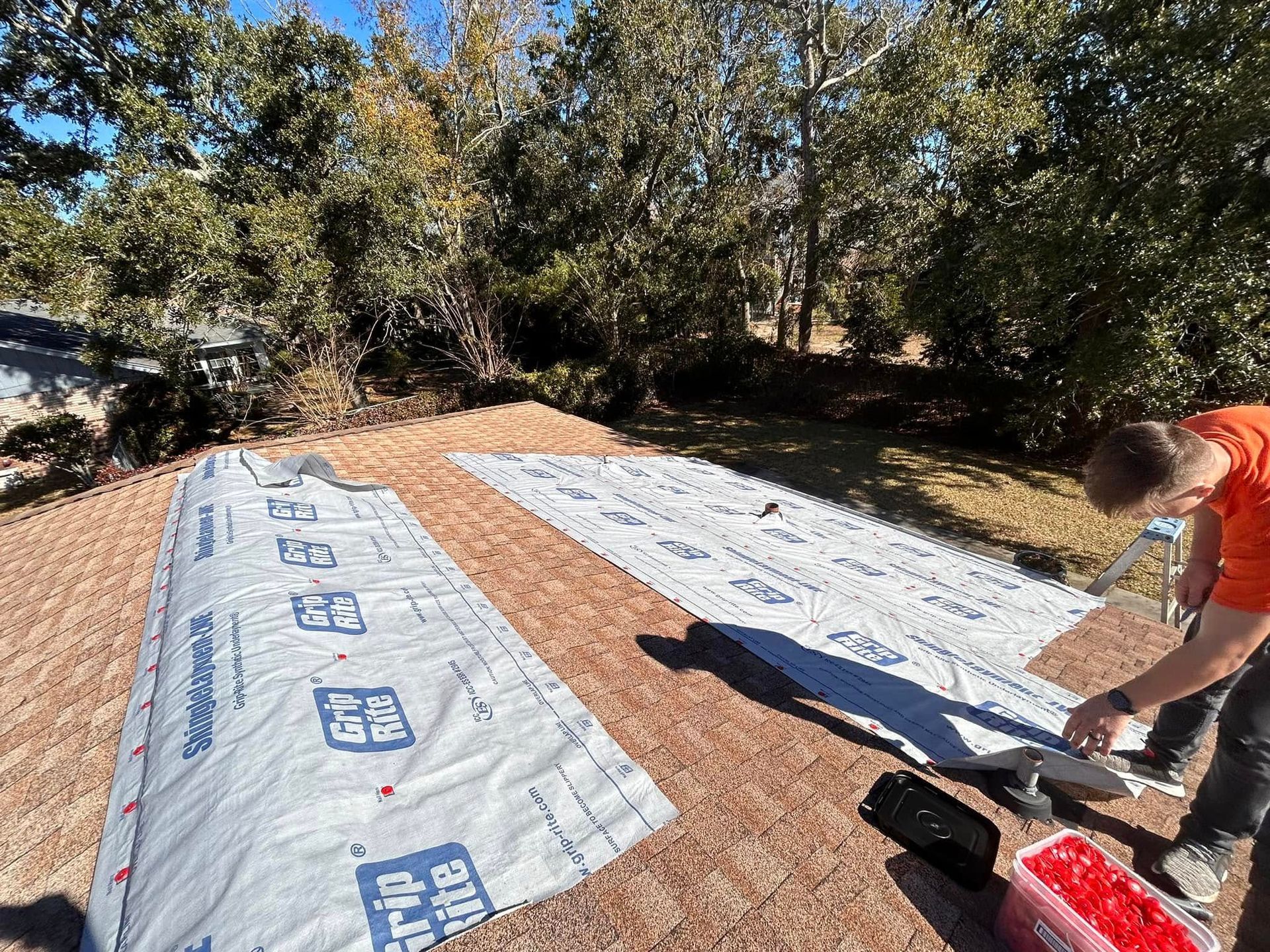 A roofer in an orange shirt installs roofing underlayment on a sunlit, wooden roof.