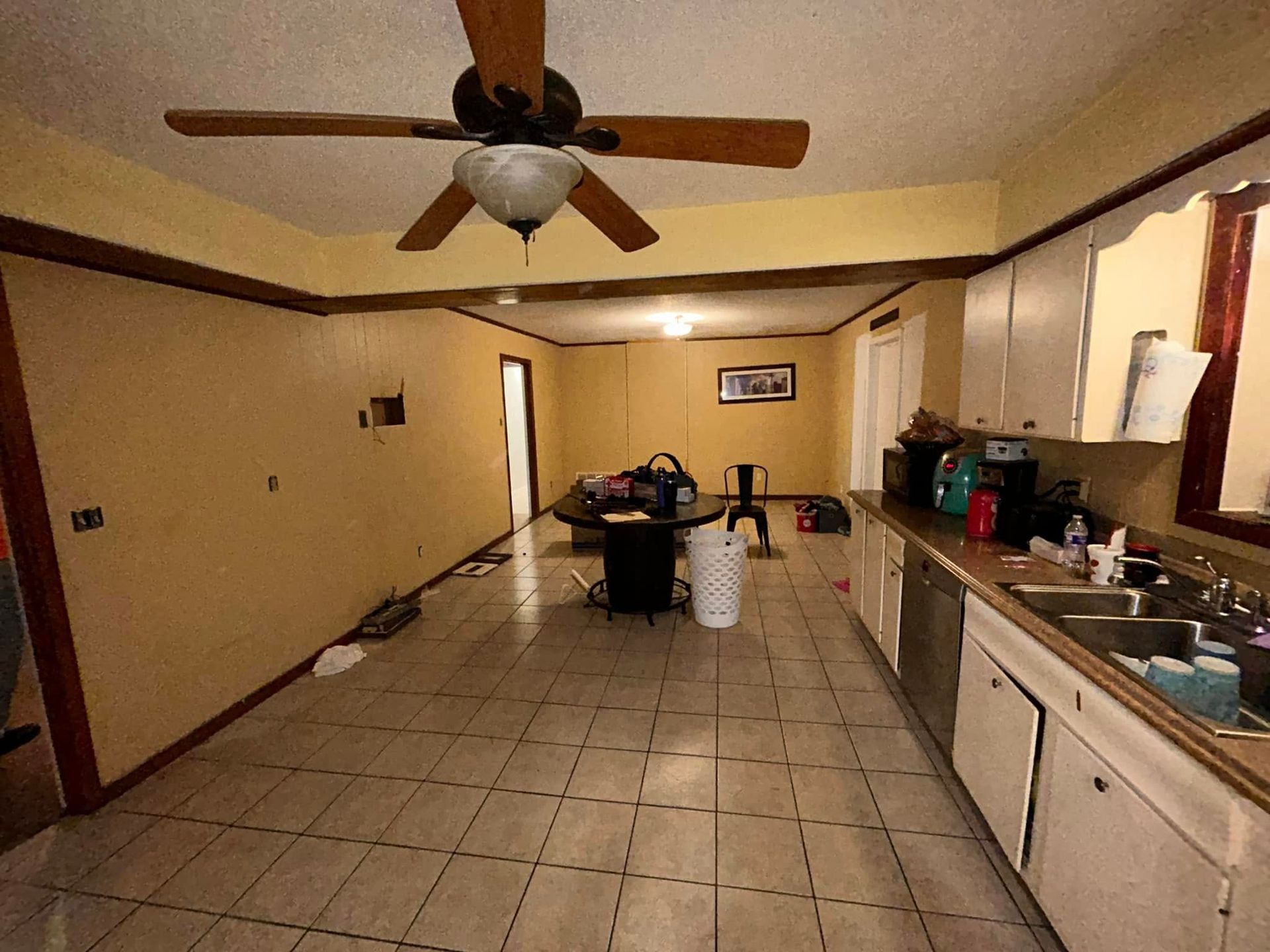 A long, slightly messy kitchen with yellow walls, a ceiling fan, and tiled floor. A table and trash can are in the center.