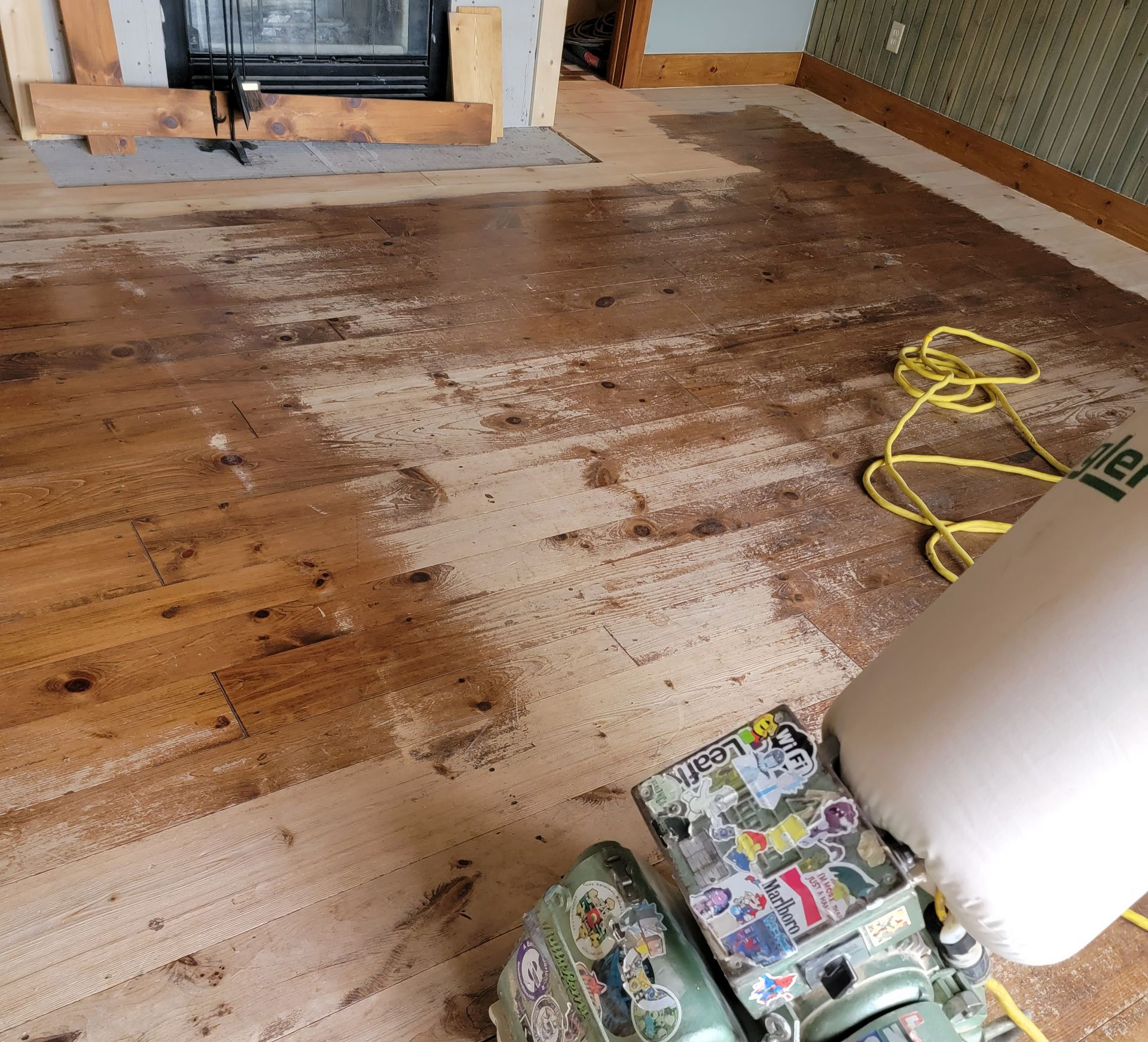 Wood floor being sanded in a room, with a floor sander and dust near a fireplace.