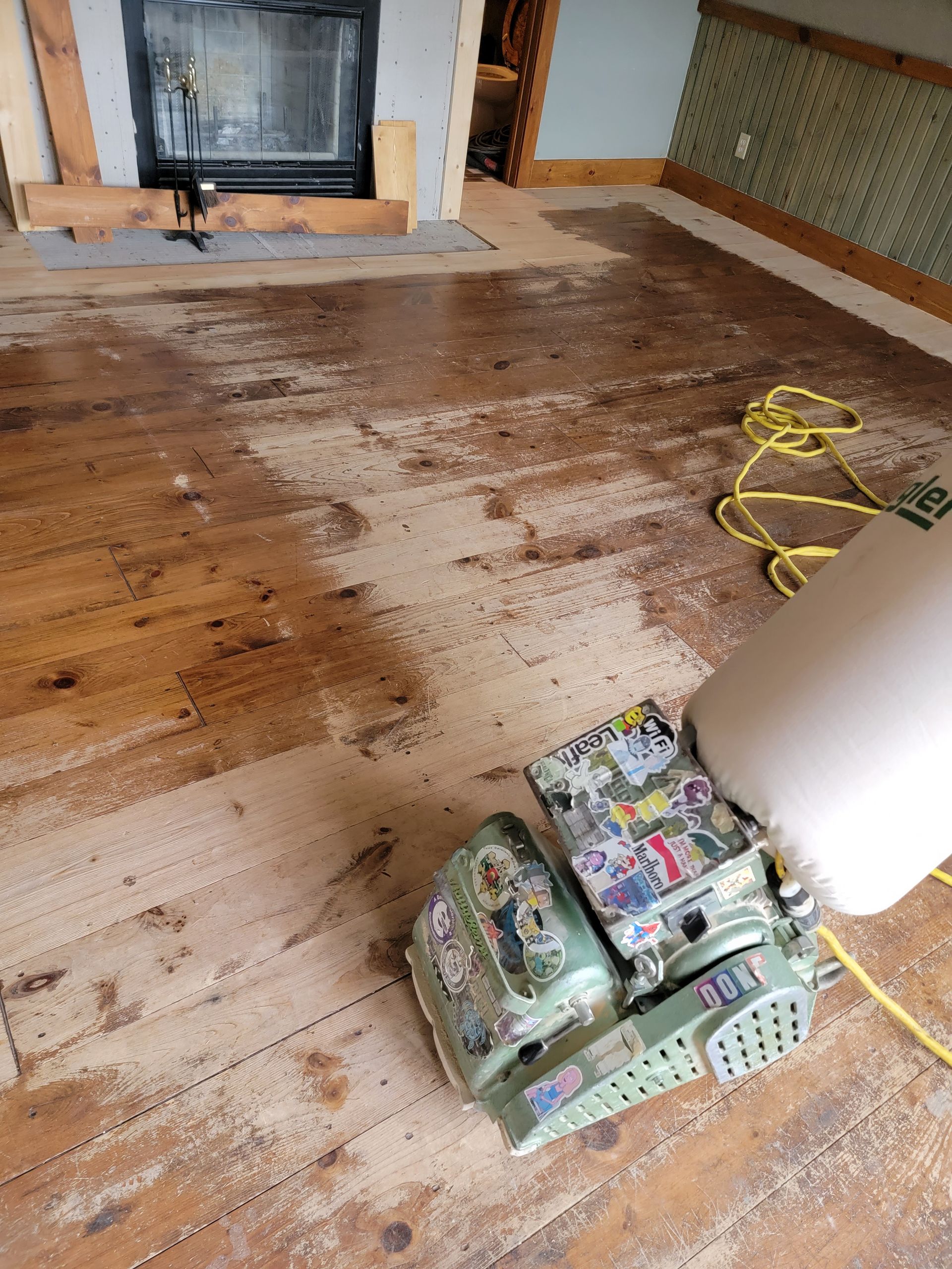 Wood floor being sanded, with a floor sander and extension cord in a room near a fireplace.