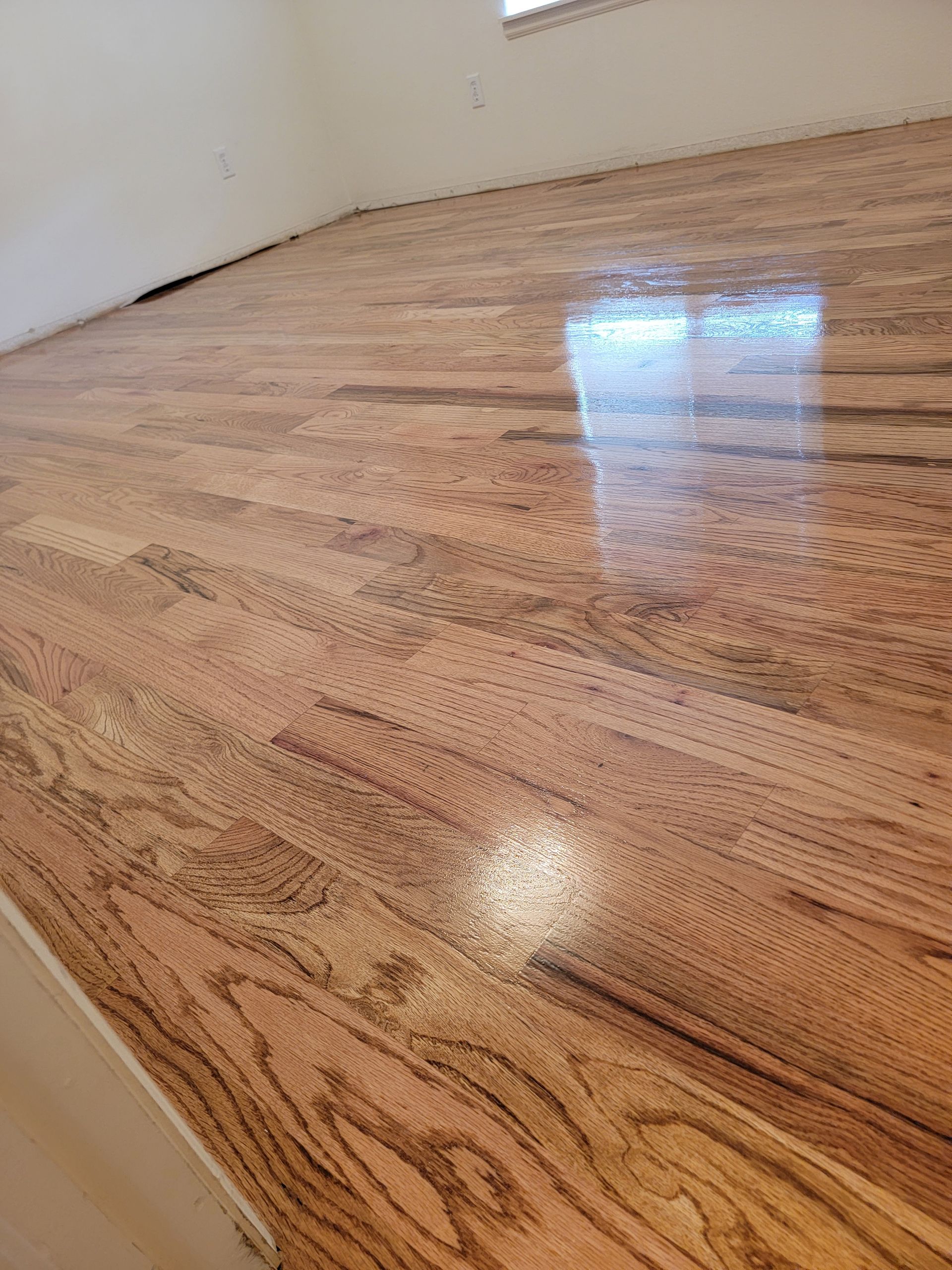 Empty room with glossy hardwood floor and white walls, reflecting light from a window