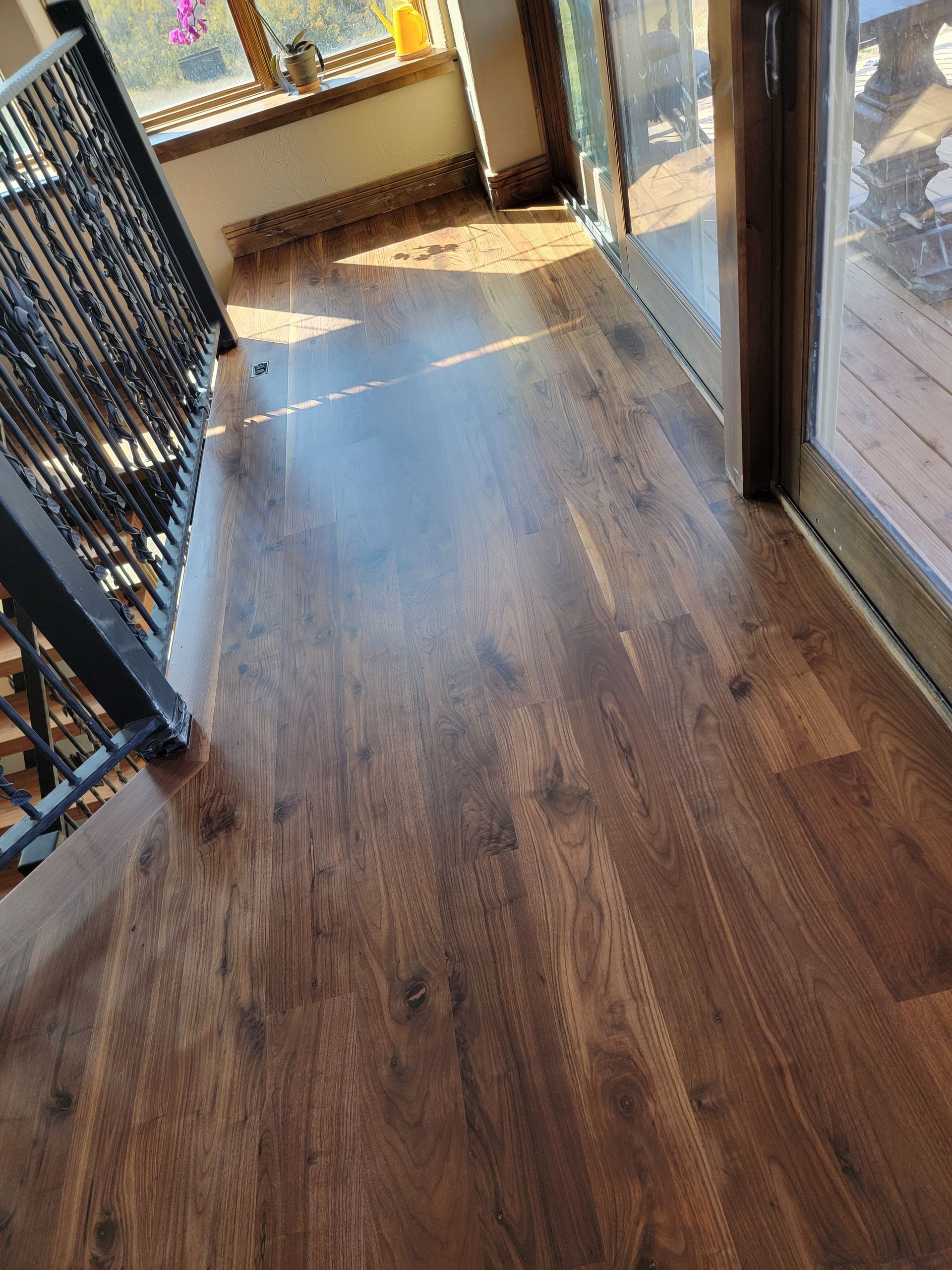 Sunlit wooden hallway with black railing, glass door, and window at the end