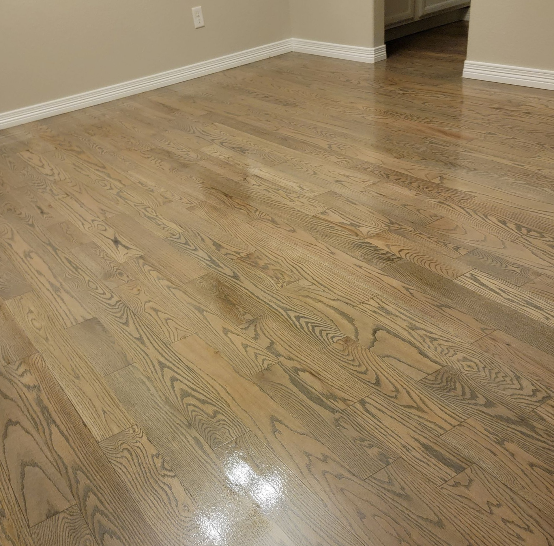 Empty room with glossy wood floor and beige walls, reflecting light near a corner window and baseboards.