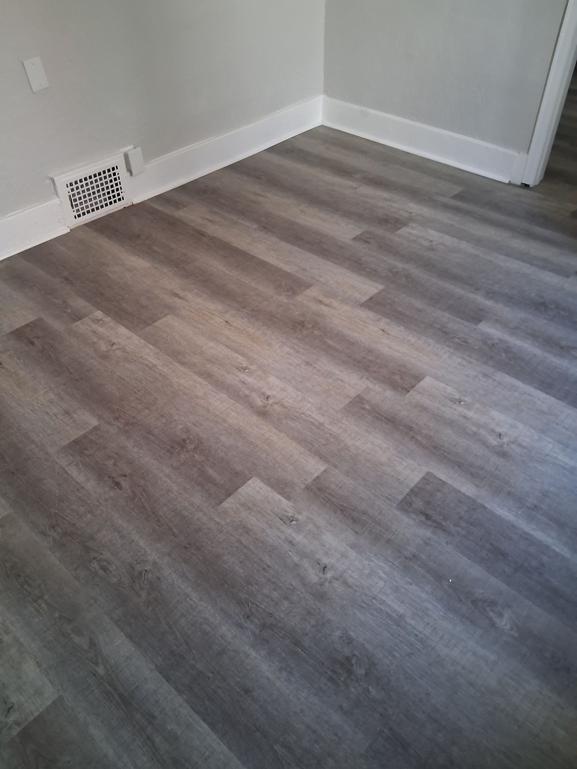 Empty room with gray wood-look flooring and white walls, showing a floor vent in the corner.