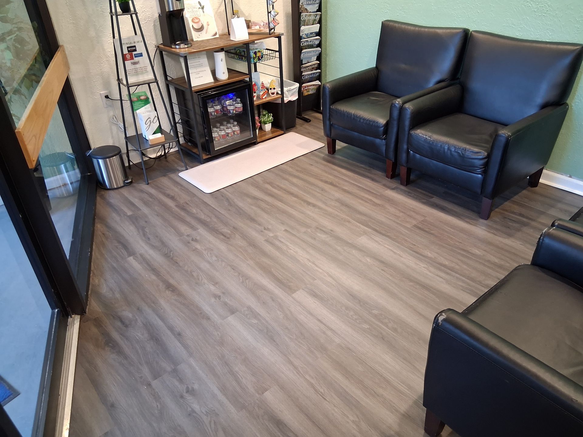 Small lobby with gray wood flooring, black leather chairs, a shelf, and a window by the entrance.