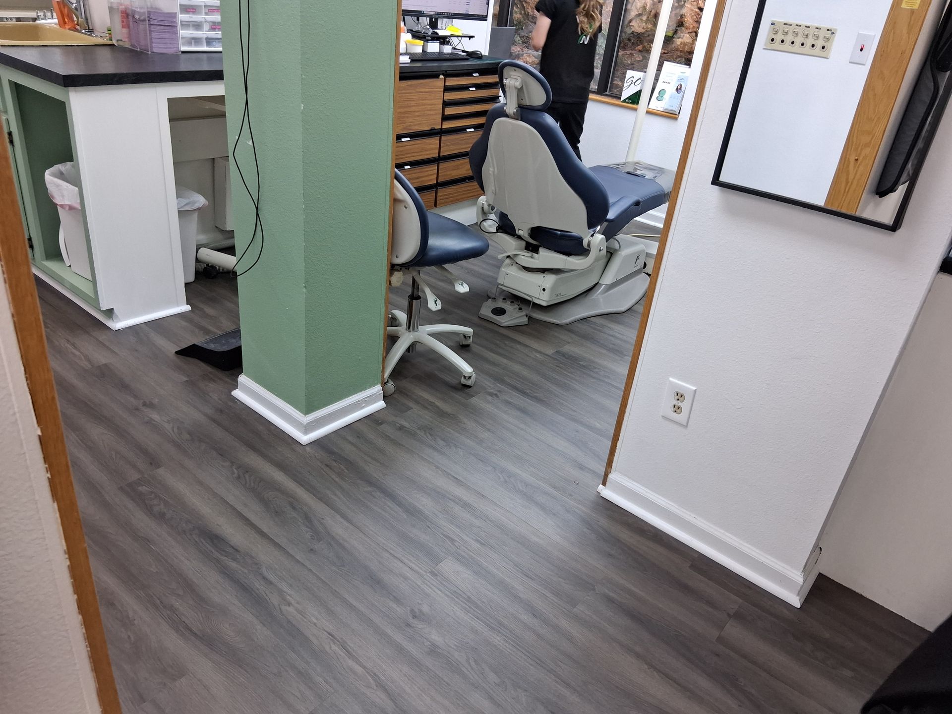 Office interior with gray flooring, green column, desks, and two white office chairs near a hallway.