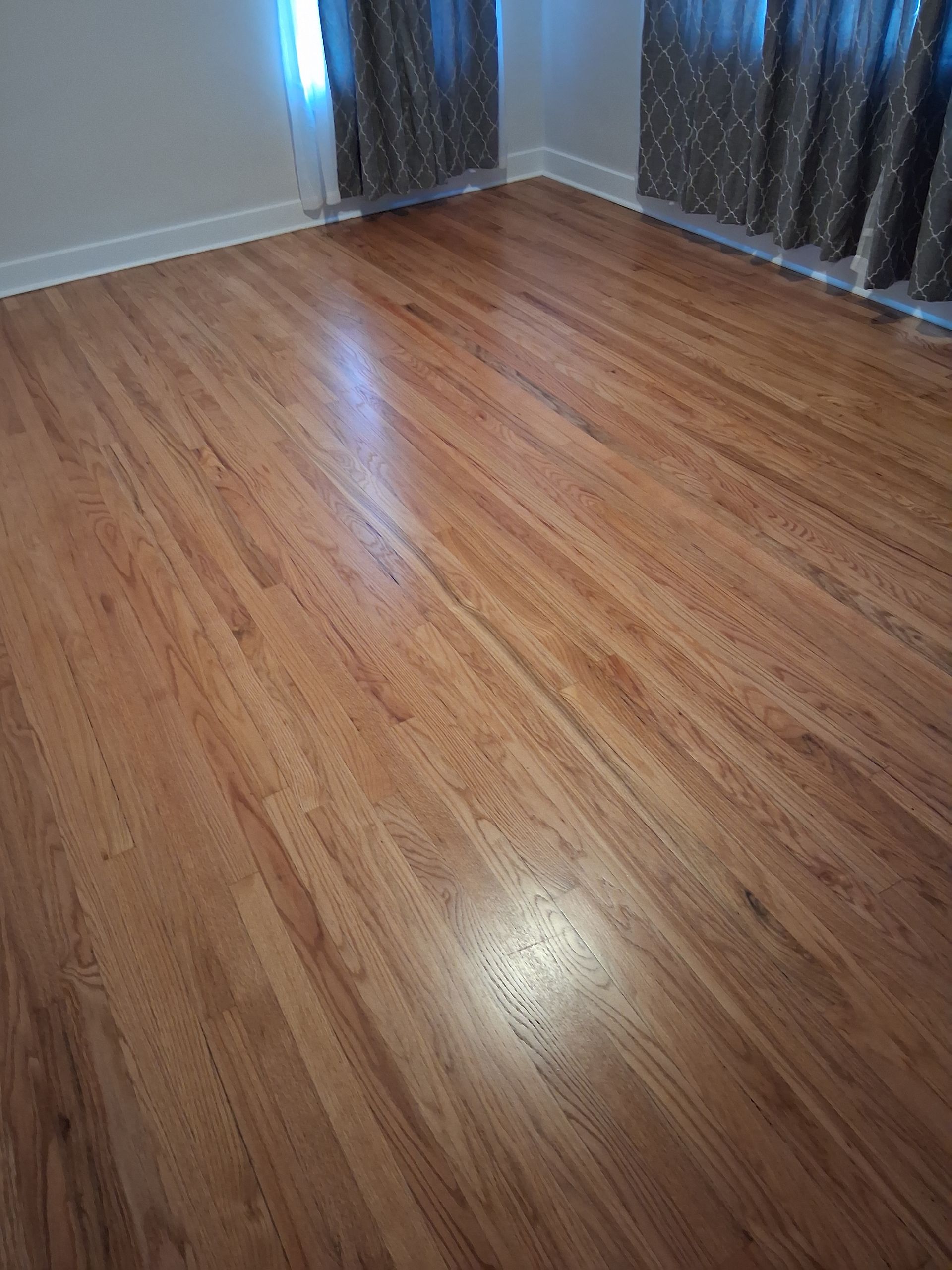 Empty room with polished hardwood floor, white walls, and dark curtains by a window.