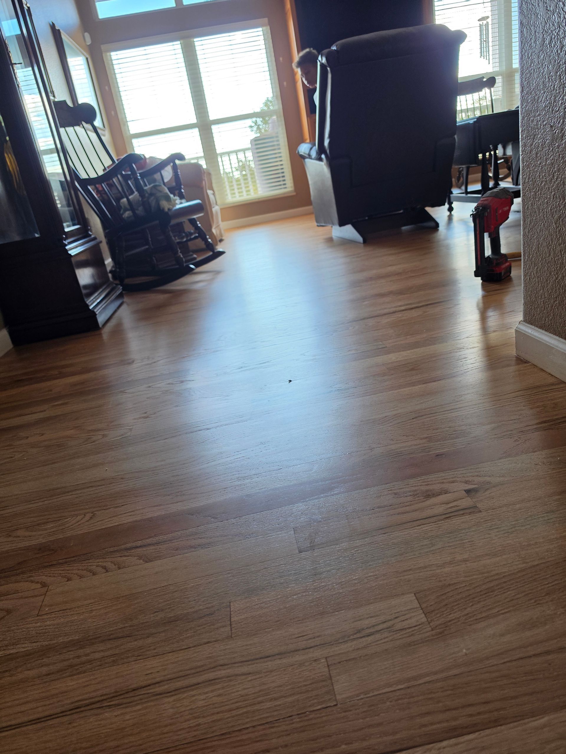 Wooden living room floor leading to a bright window, with chairs and furniture in the background.
