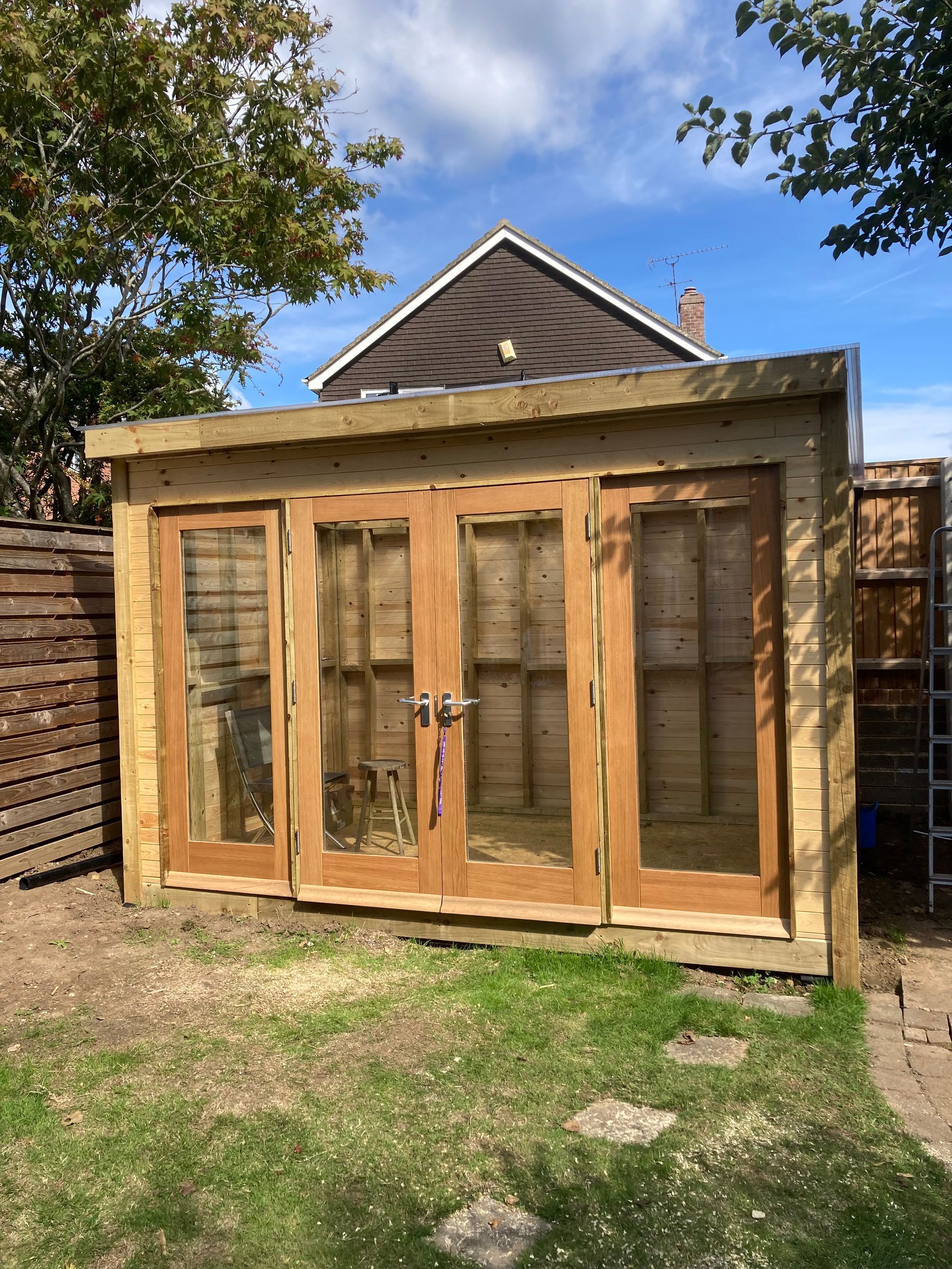 Wooden garden shed with glass double doors and windows in a grassy backyard.