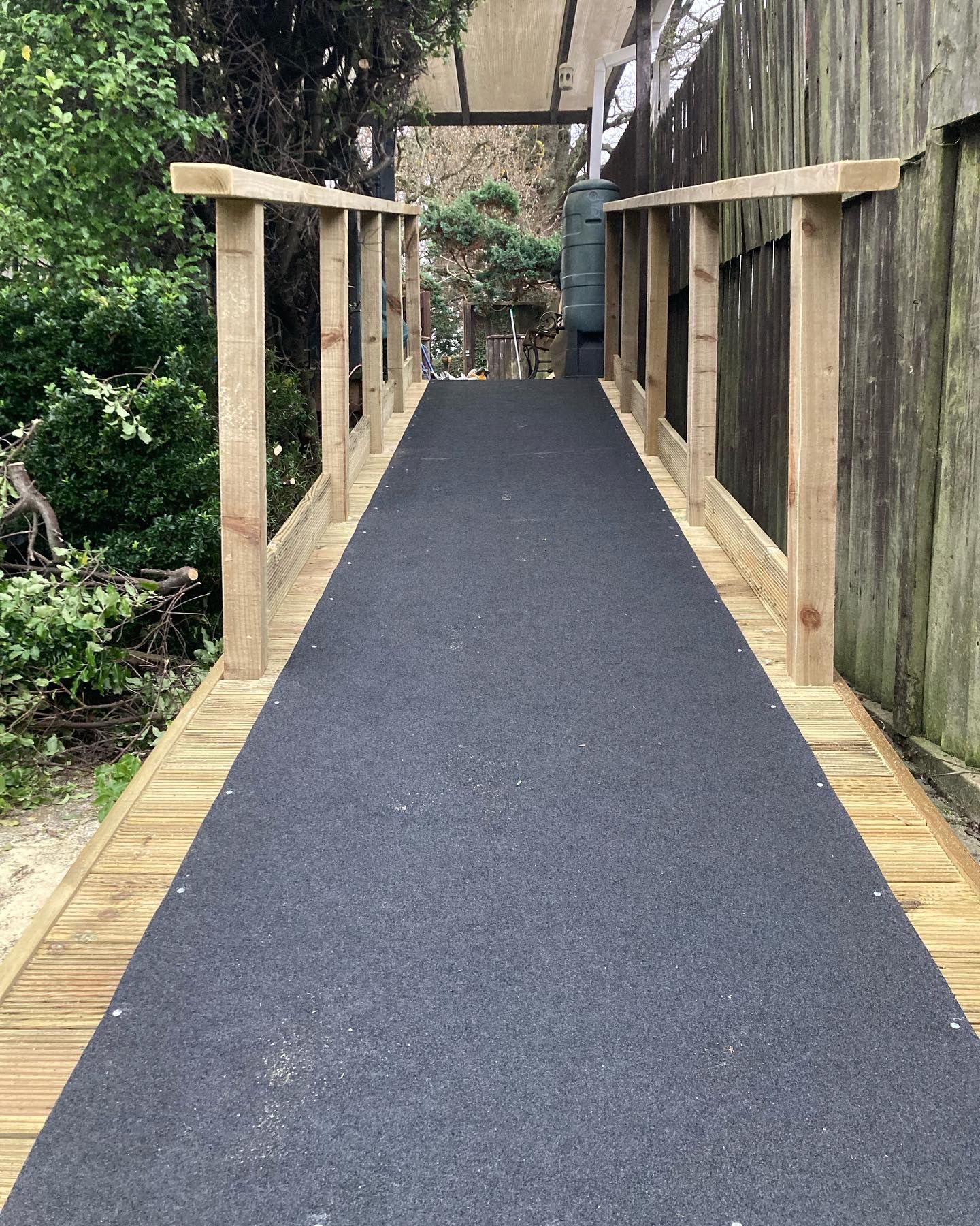 Narrow outdoor wooden walkway with black paved surface and railings through a garden path