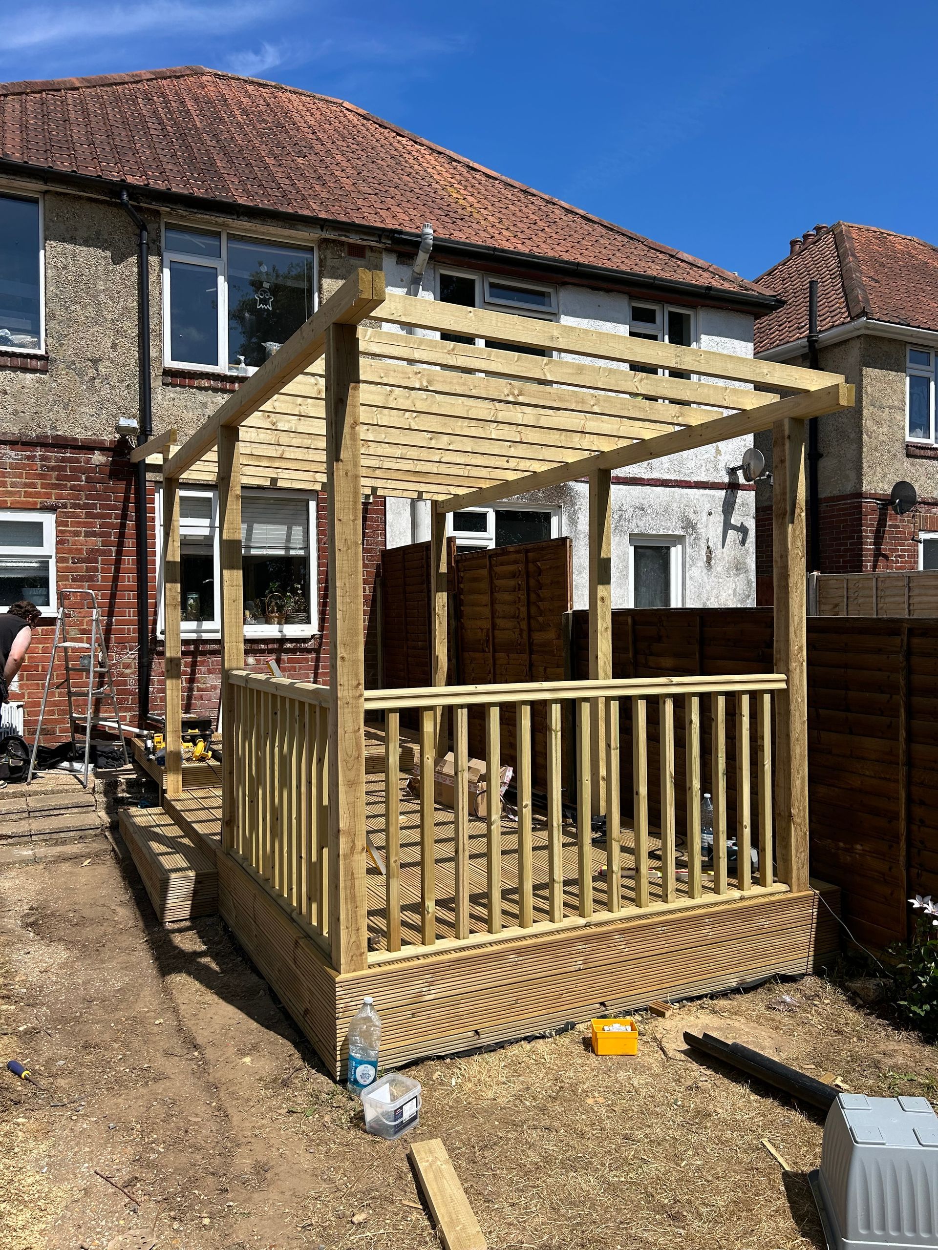 Wooden gazebo frame with slatted railings in a gravel backyard beside a brick house.