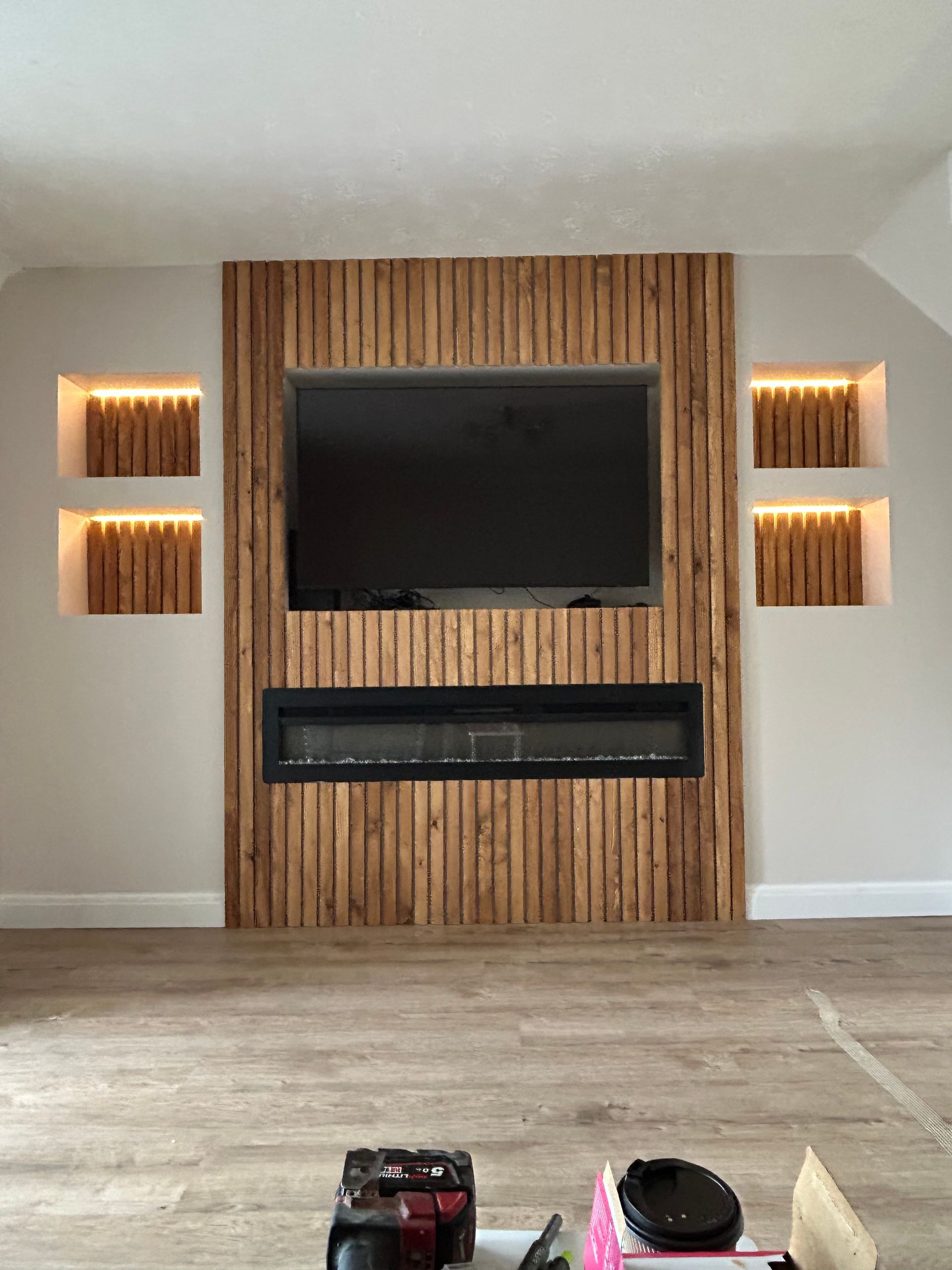 Modern living room with a wood-paneled fireplace wall, wall-mounted TV, and lit recessed shelves.