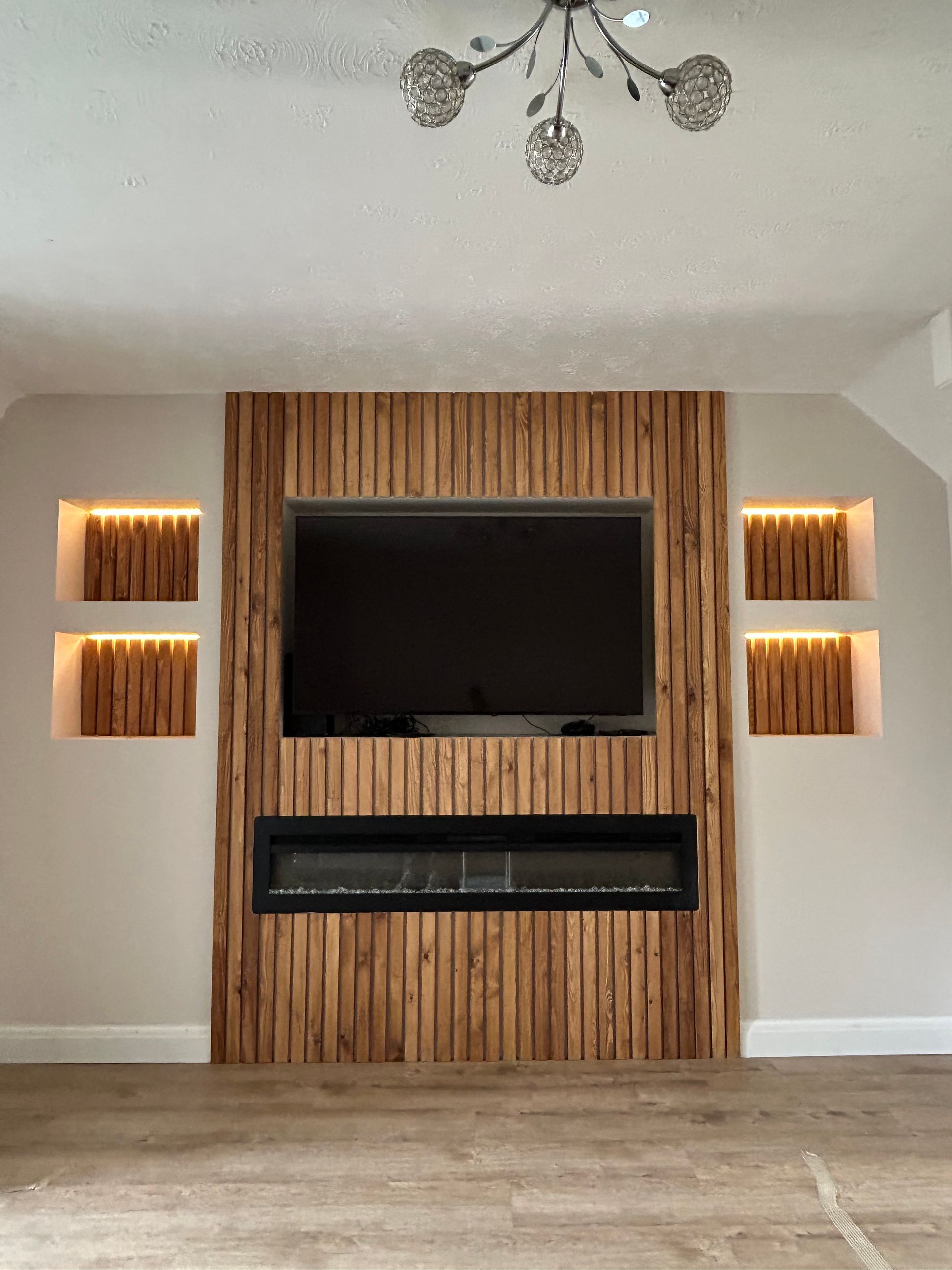 Living room with wall-mounted TV and electric fireplace on a vertical wood accent wall, flanked by lit shelves