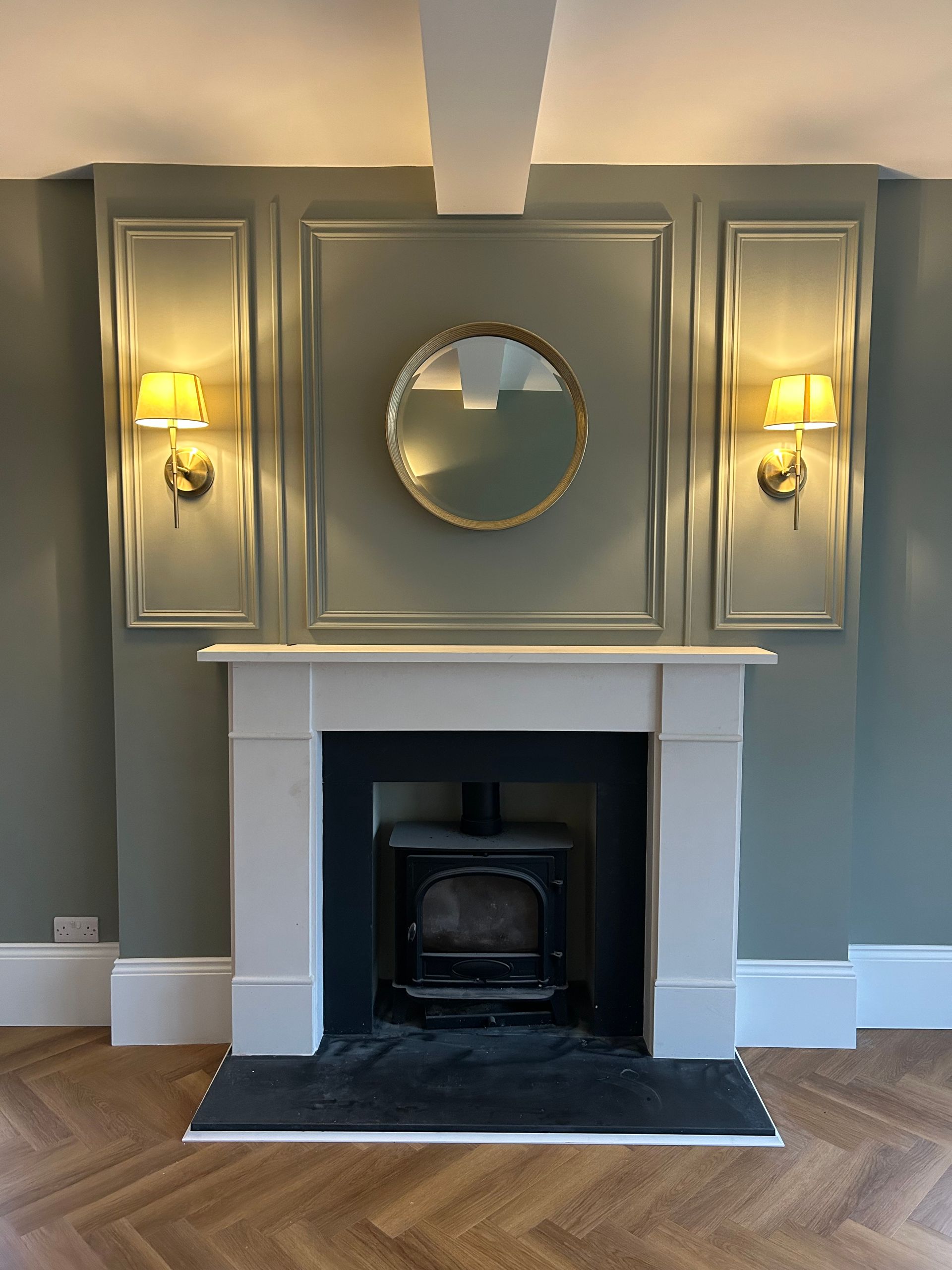 Gray living room fireplace with white mantel, round mirror, wall sconces, and herringbone wood floor.