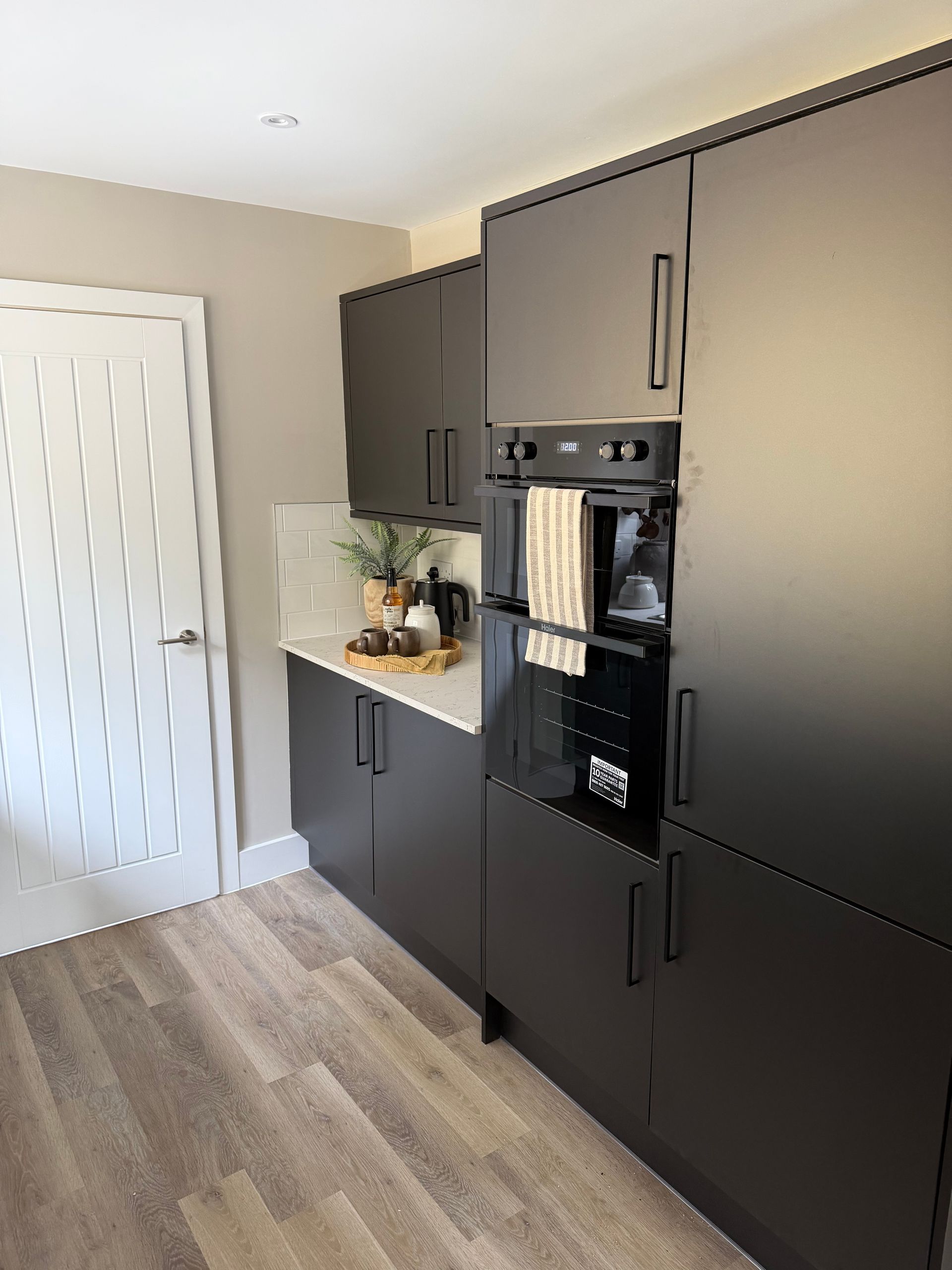 Modern kitchen with dark cabinets, built-in oven, countertop decor, and a white door on the left.