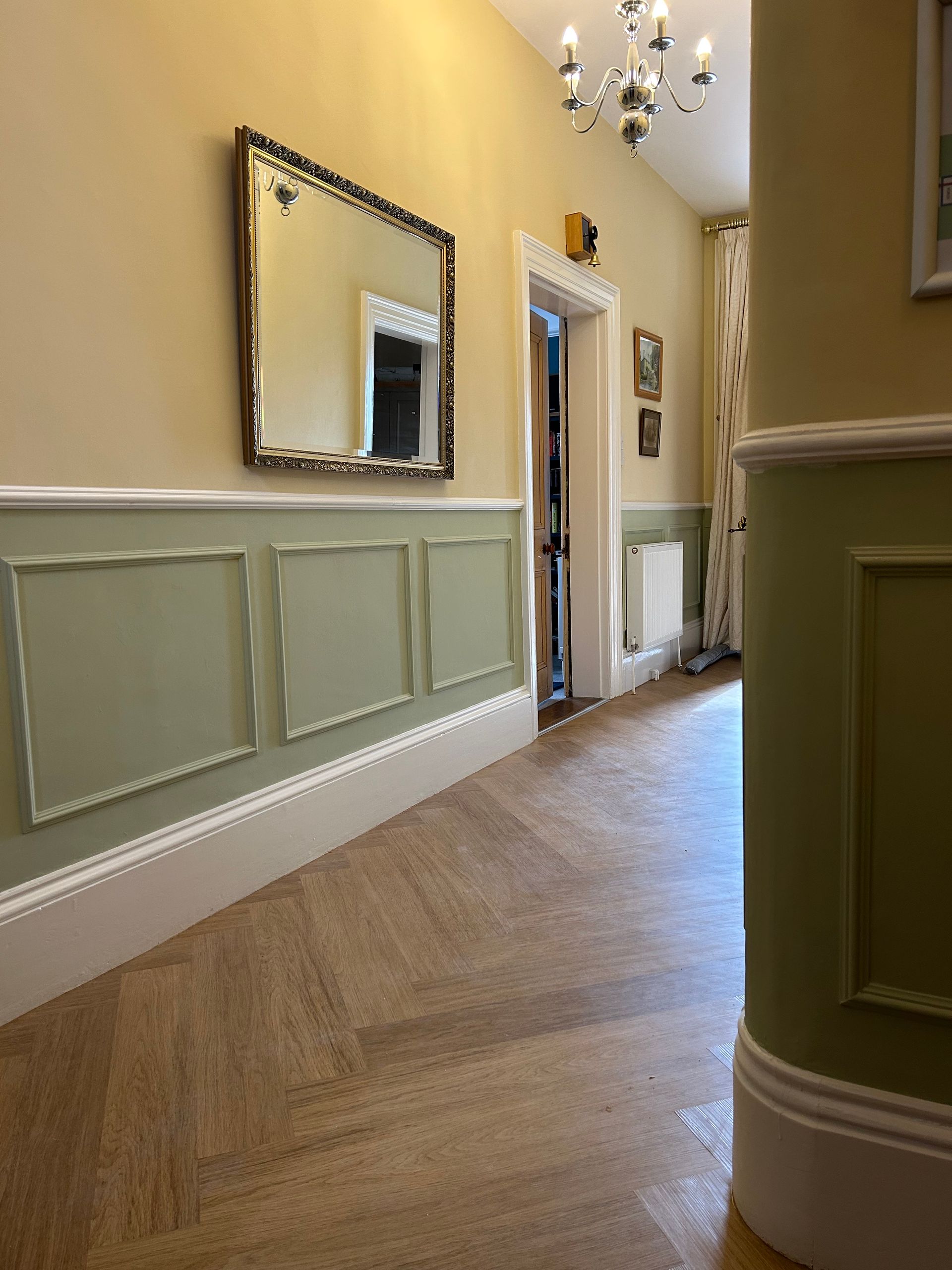 Hallway with green wainscoting, yellow walls, mirror, chandelier, and hardwood floor leading to a doorway