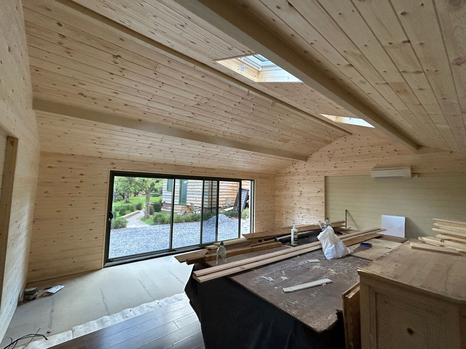 Wood-paneled room under construction with skylights, large windows, and building materials stacked inside.