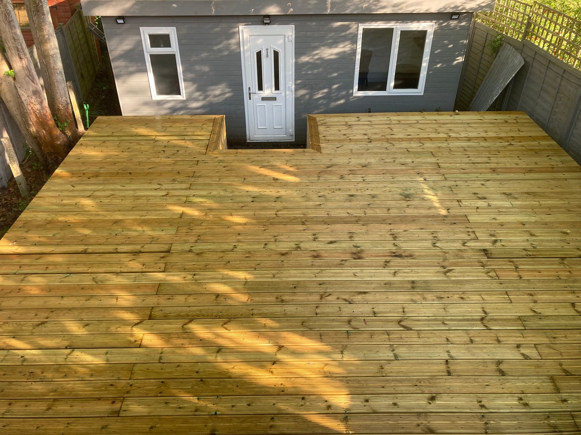 Wooden backyard deck with a gray shed wall, white door and windows, lit by sunlight and tree shadows