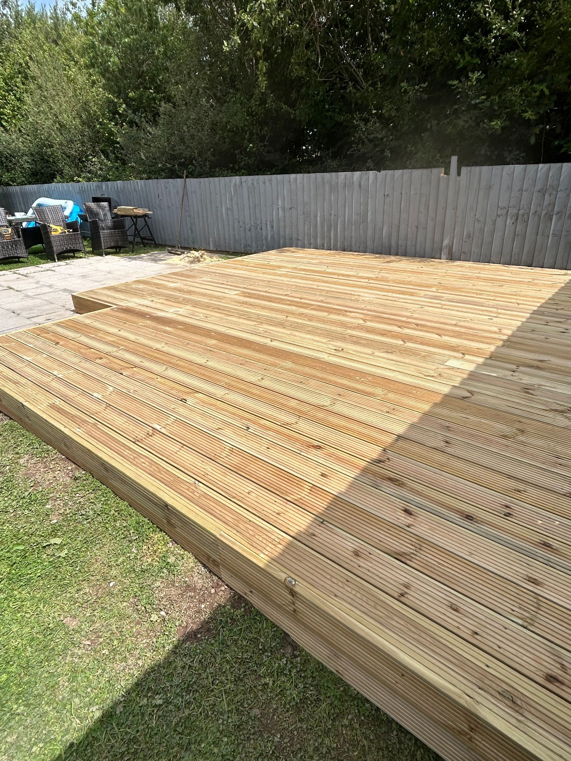 Stacked wooden planks in a backyard beside a fence and grass, with bins and trees in the background.