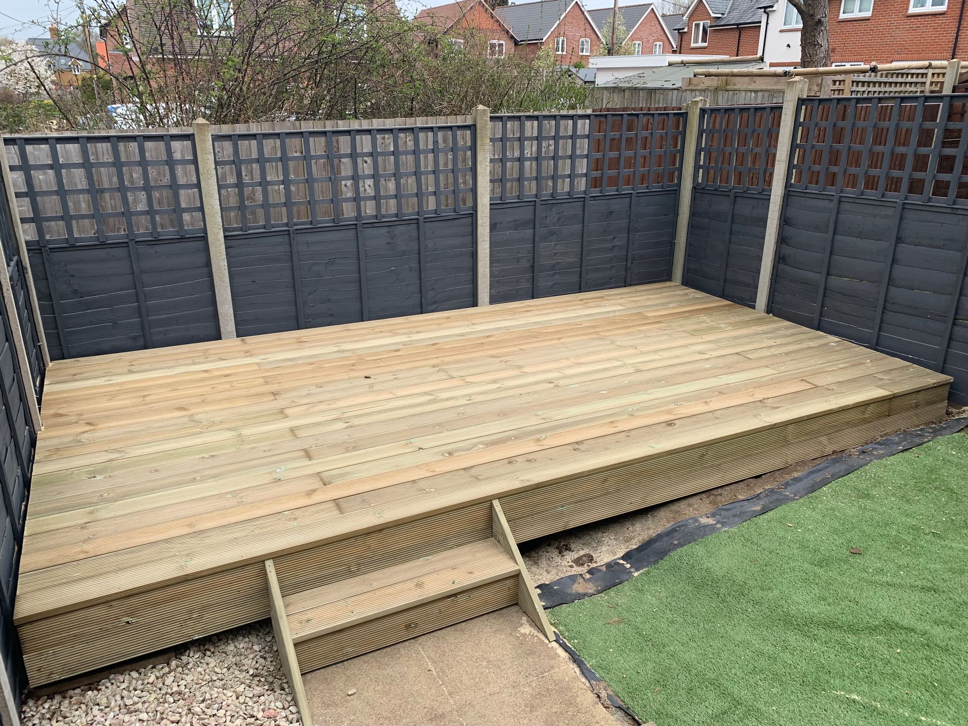 Wooden backyard deck with steps, dark fence panels, and green lawn beside it.