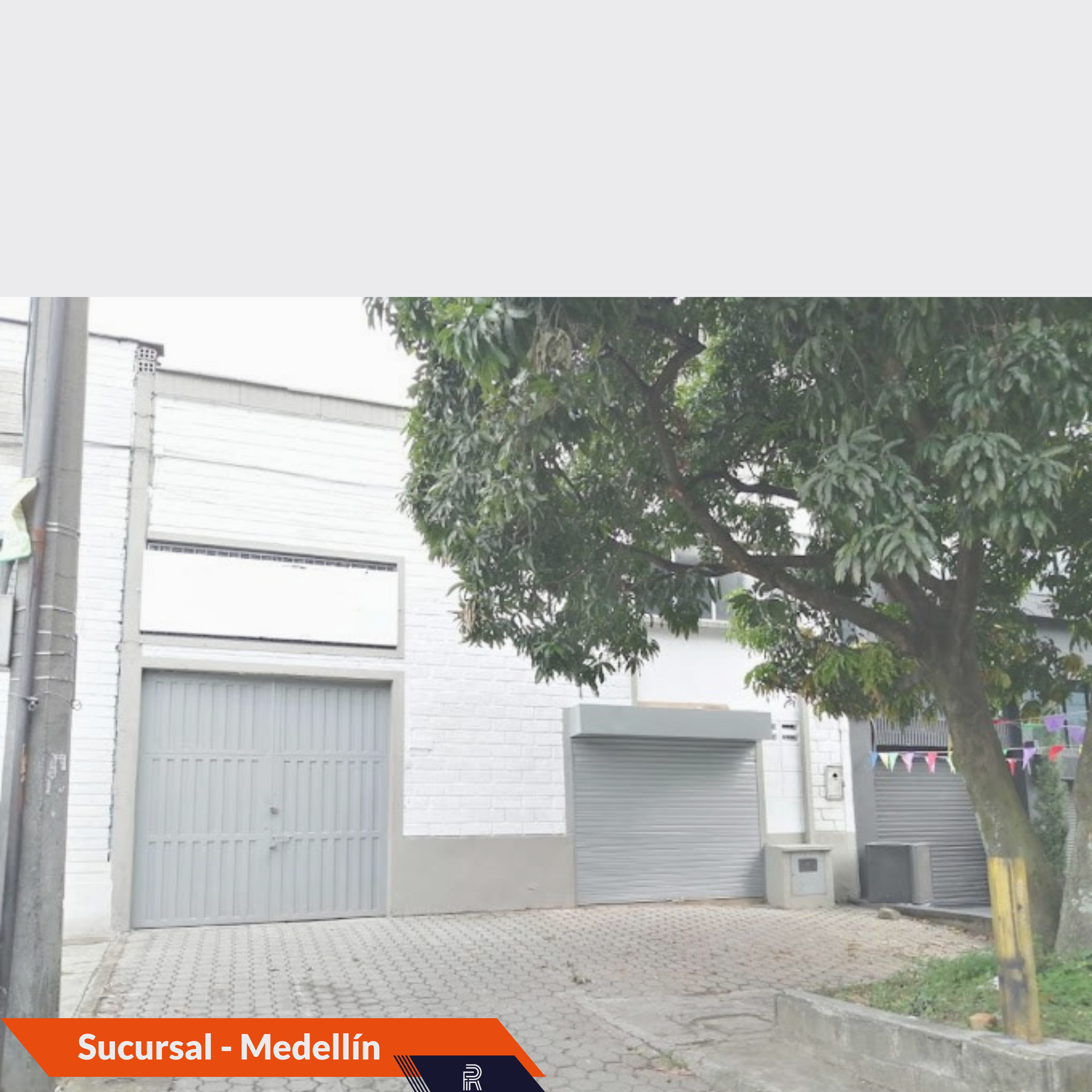 Almacén en Medellín con dos puertas de garaje cerradas. Un árbol a la derecha.