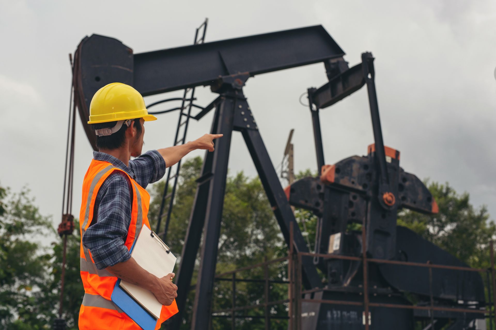 Un trabajador con chaleco de seguridad y casco señala una bomba de extracción de petróleo en un día nublado.