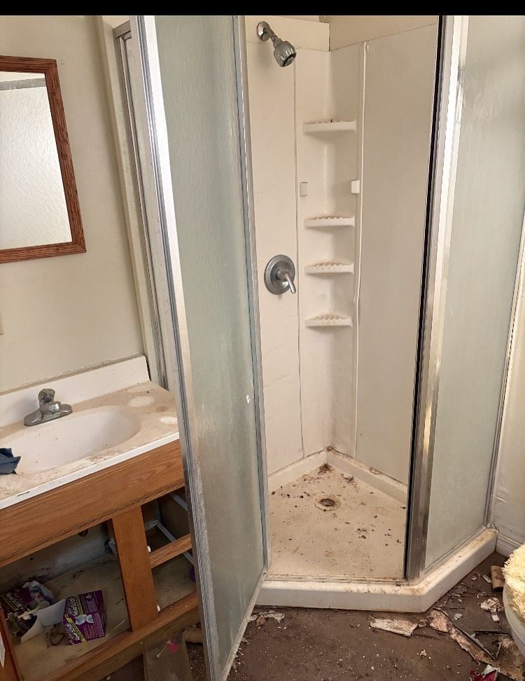 Bathroom with a white corner shower, sink, and mirror. The room appears dirty and in need of repair.
