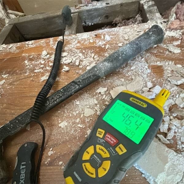 A moisture meter reading 46.4% on a wood surface with a pry bar in a dusty, indoor setting.