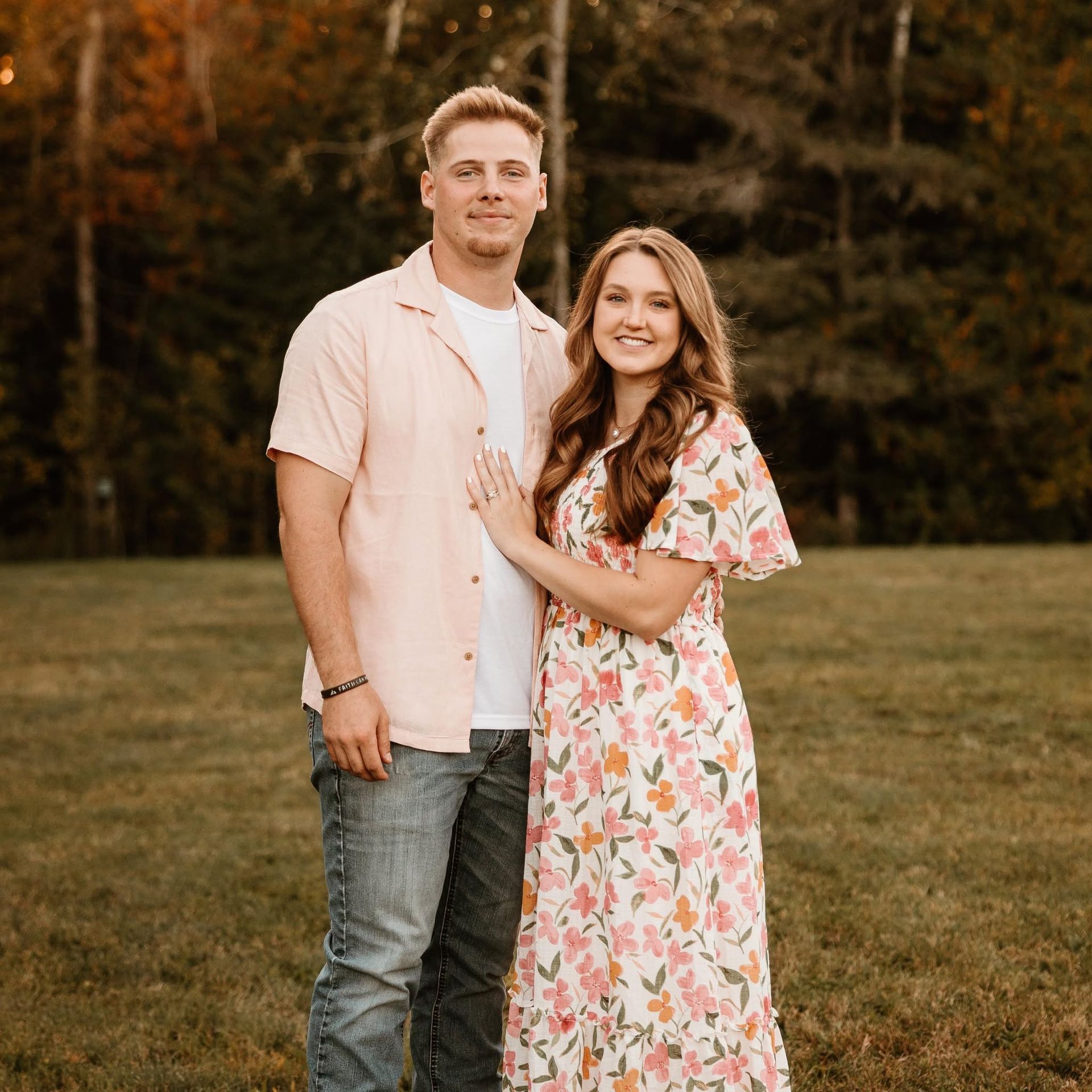 Couple standing in a grassy field, smiling, with autumn trees in the background.