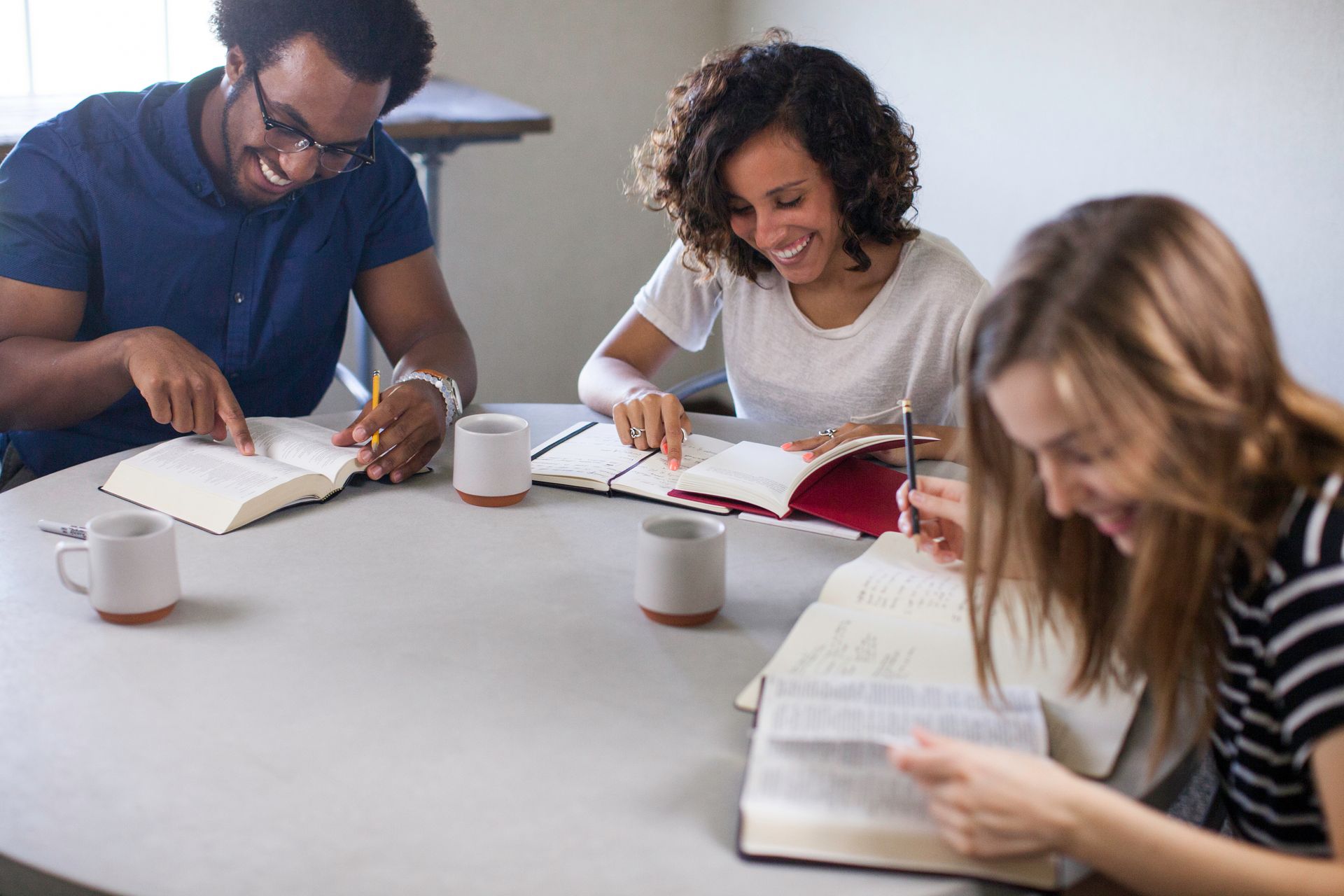 Three people reading and writing at a table in a bright classroom or study room