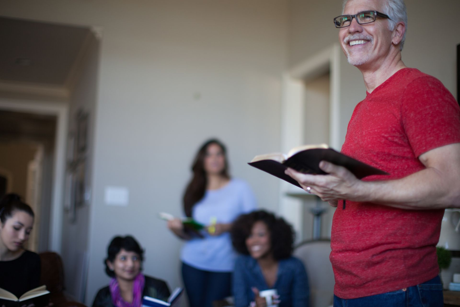 Smiling man in red shirt reading from an open book to a small seated group indoors