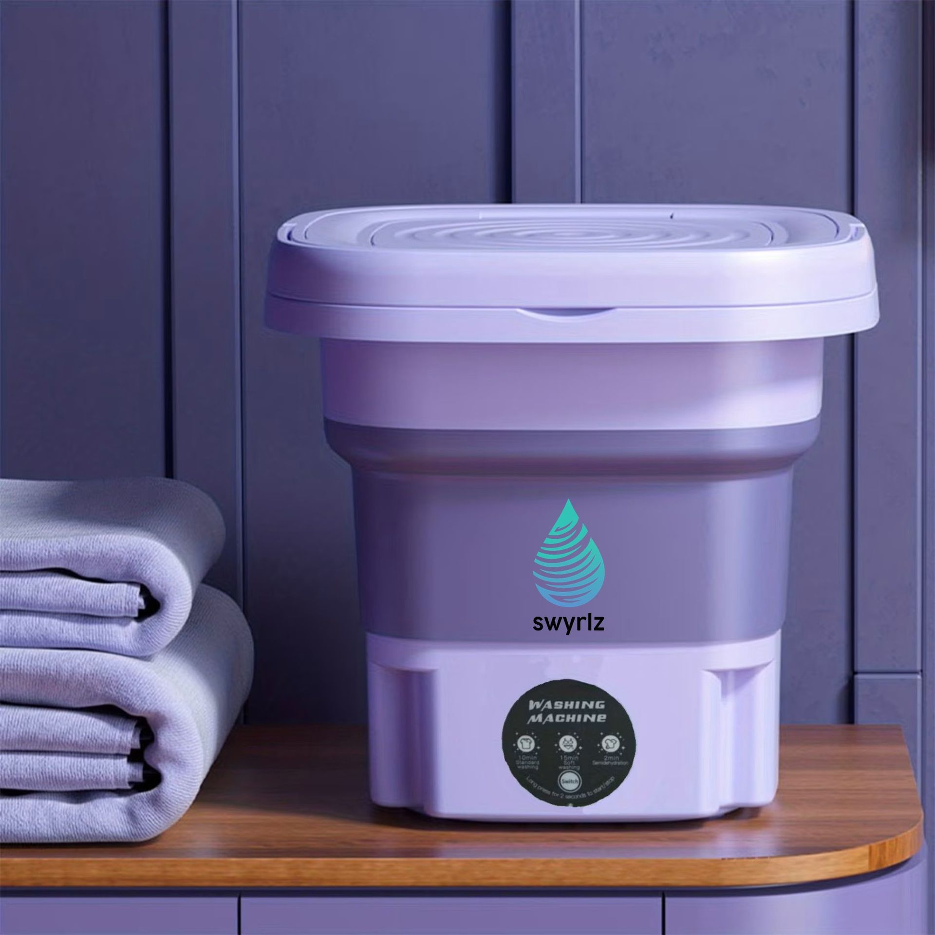 Collapsible mini washing machine, purple, on a wooden surface, next to folded towels.